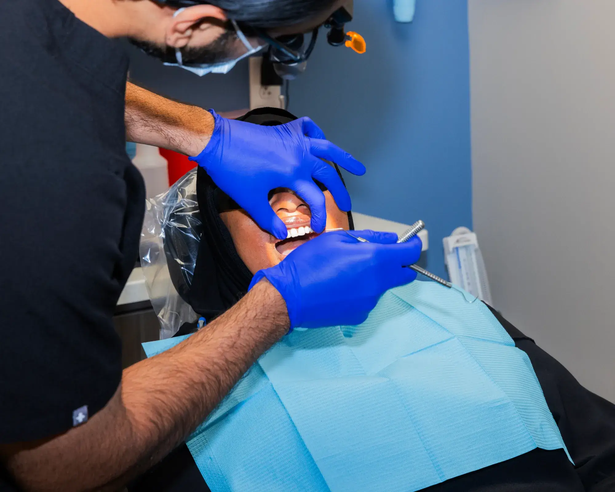 A dentist wearing blue gloves examines a patient's teeth using a dental mirror.