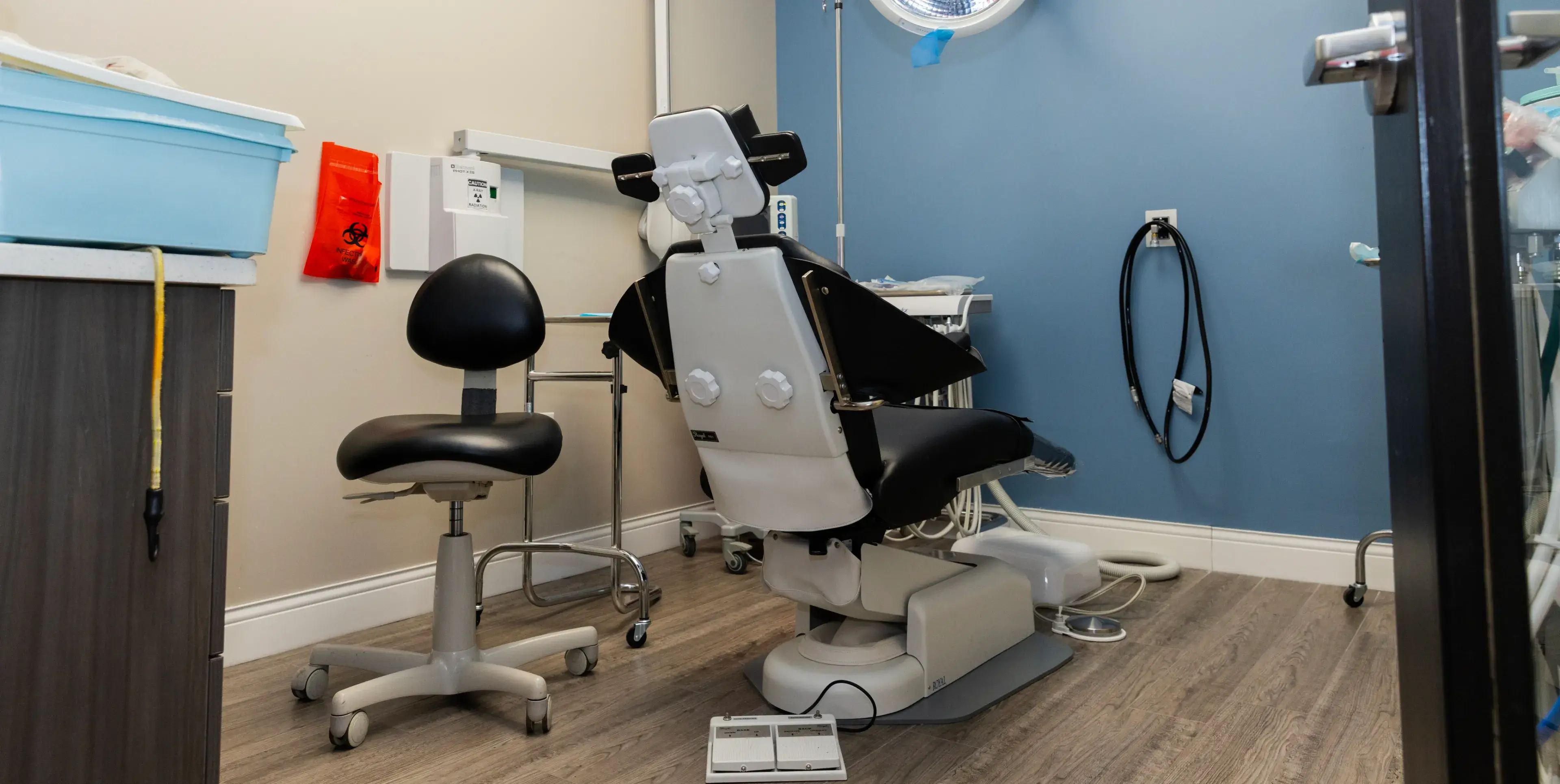 Dental examination room with a dentist chair, stool, and medical equipment on blue and beige walls.