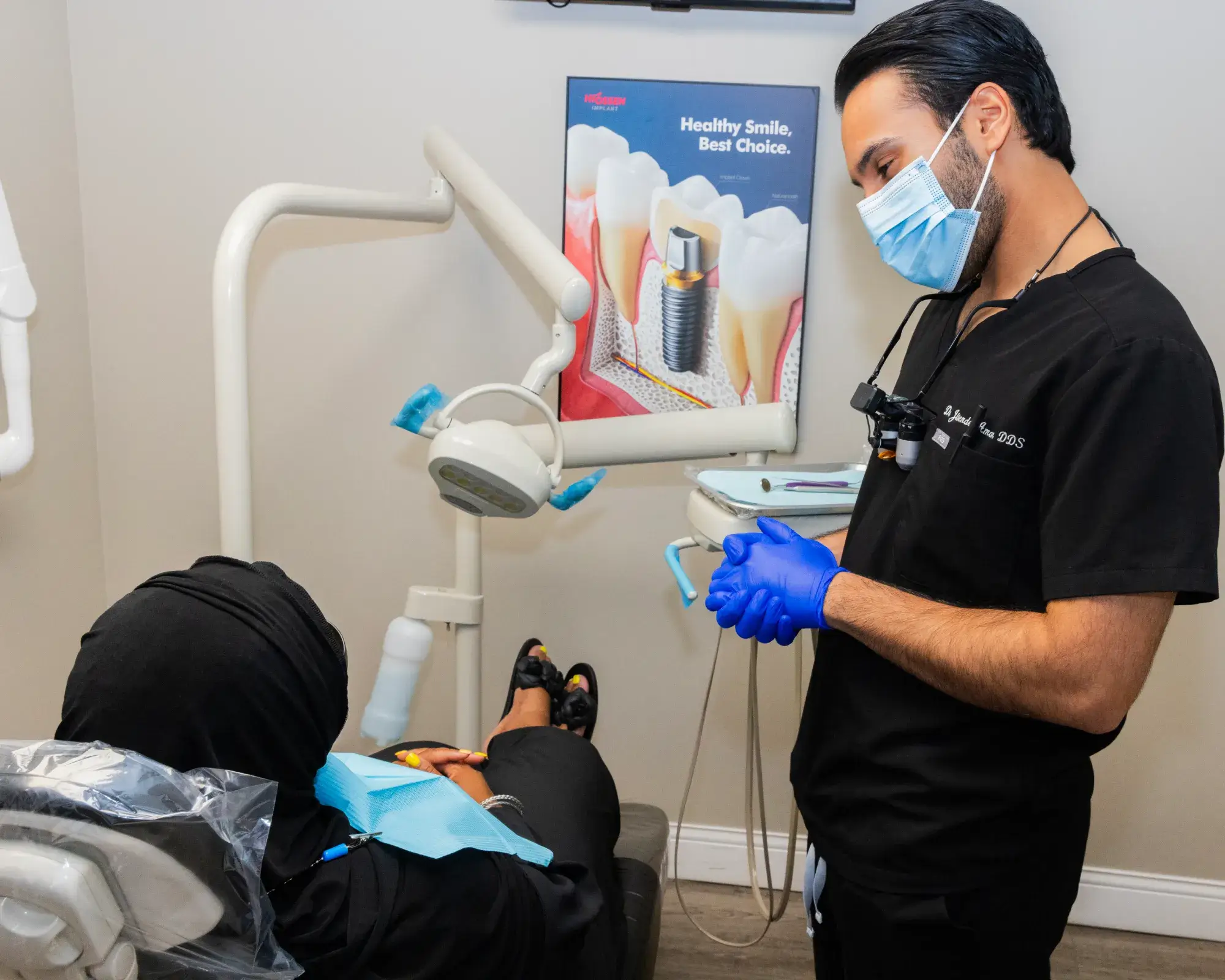 A dentist in a mask and gloves talks to a patient sitting in a dental chair.