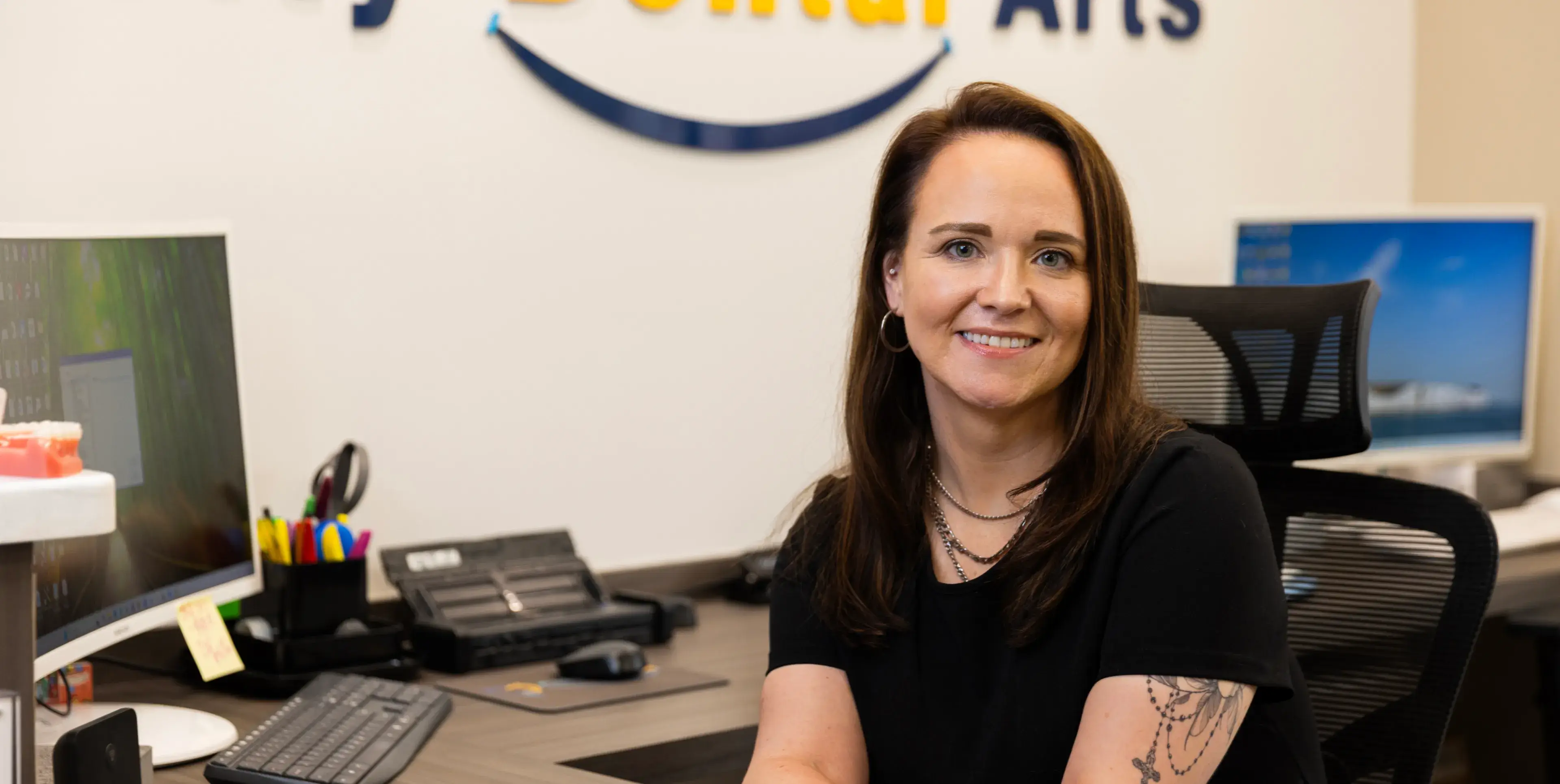 A woman with long hair and a tattooed arm sits at an office desk, smiling.