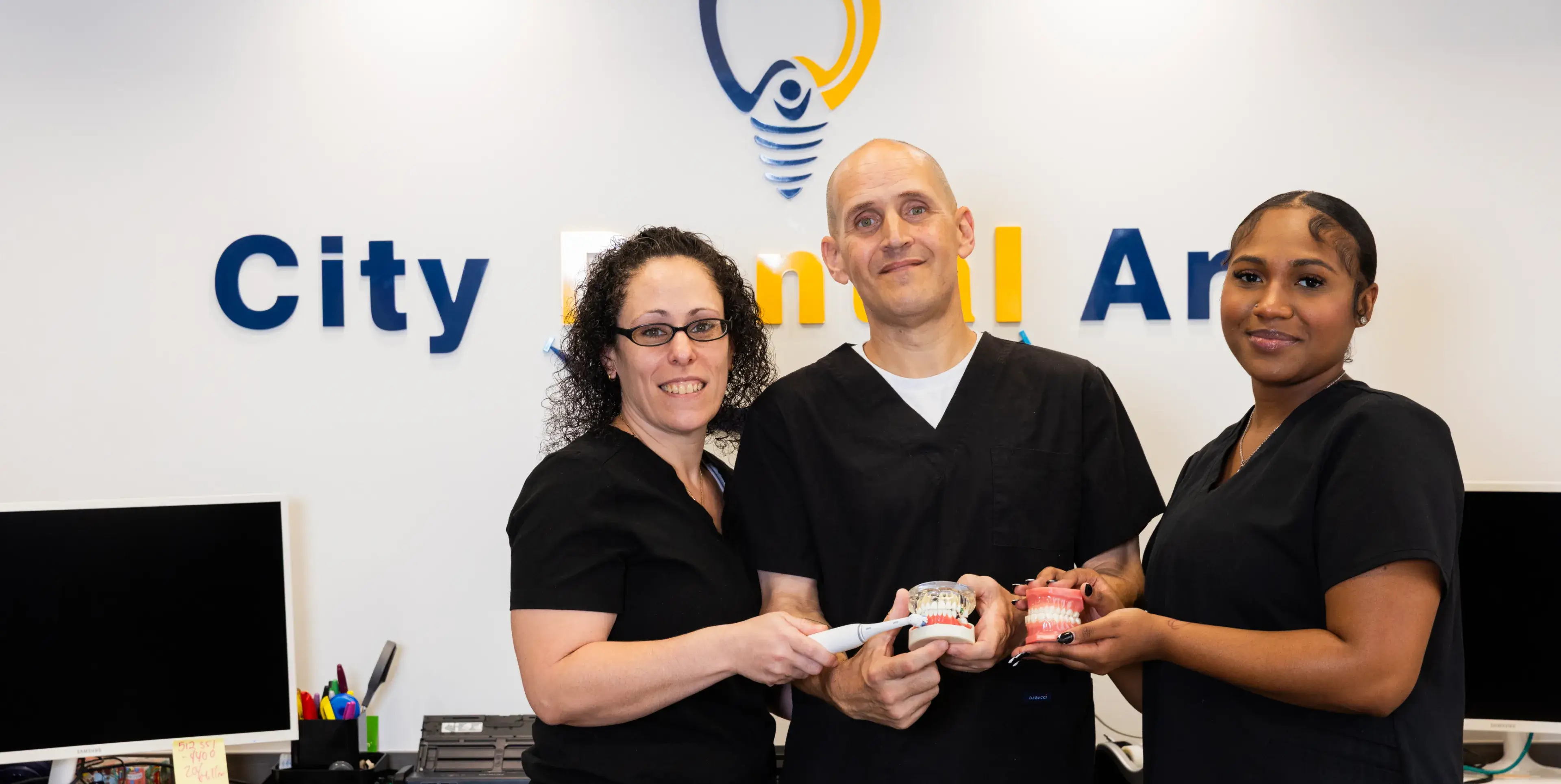 Three dental professionals holding dental models and a toothbrush, smiling in front of a dental office sign.