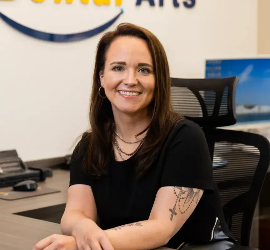 A woman with brown hair sitting at an office desk, smiling at the camera.