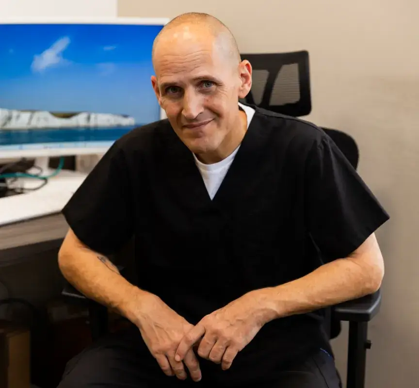 A person in black scrubs sits in an office chair, smiling at the camera.