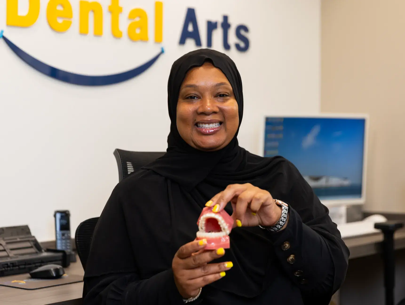 A woman in a black headscarf holds dental model teeth in a dental office.