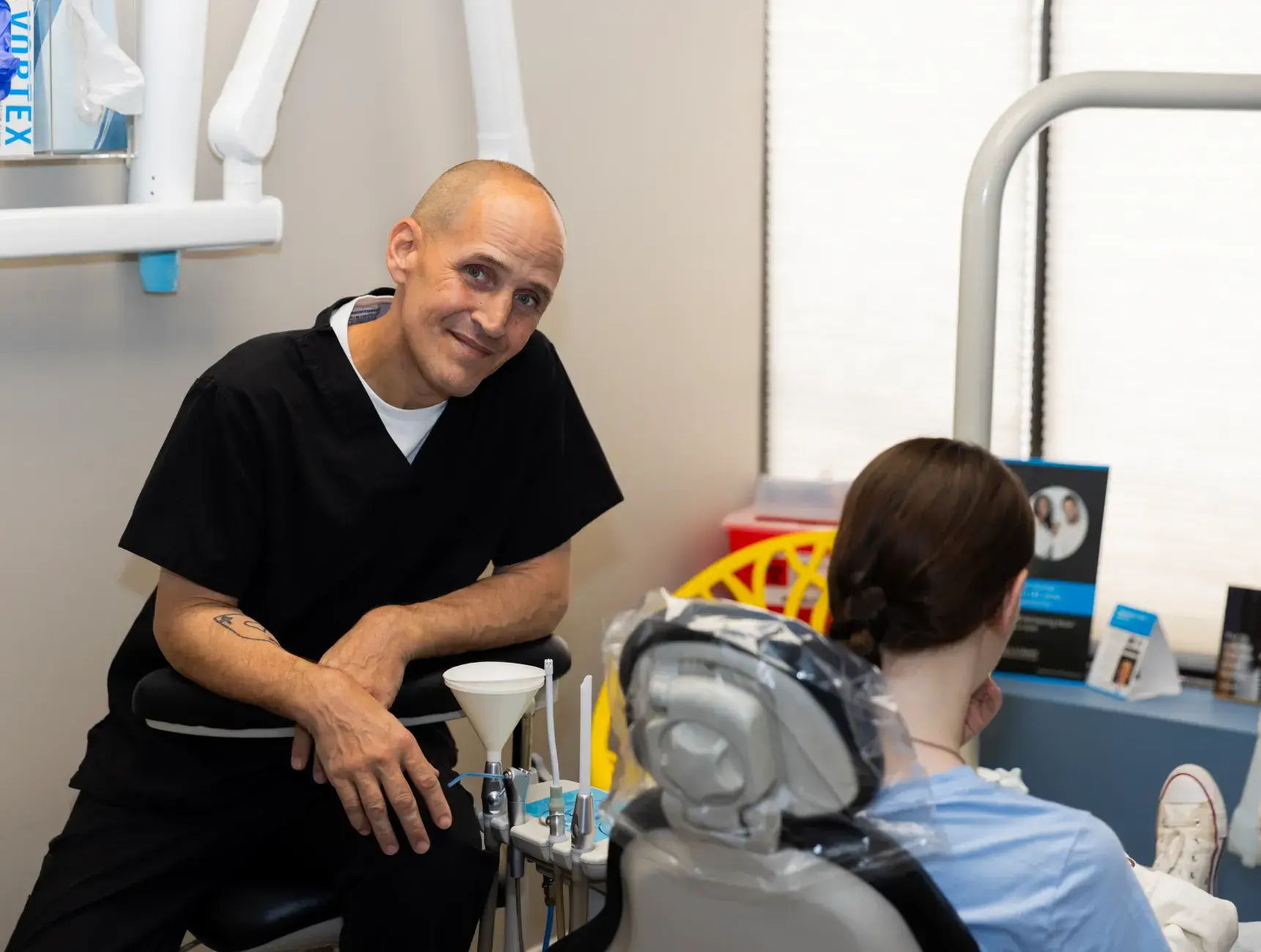 A dentist in black scrubs smiles at a seated patient in a dental clinic.
