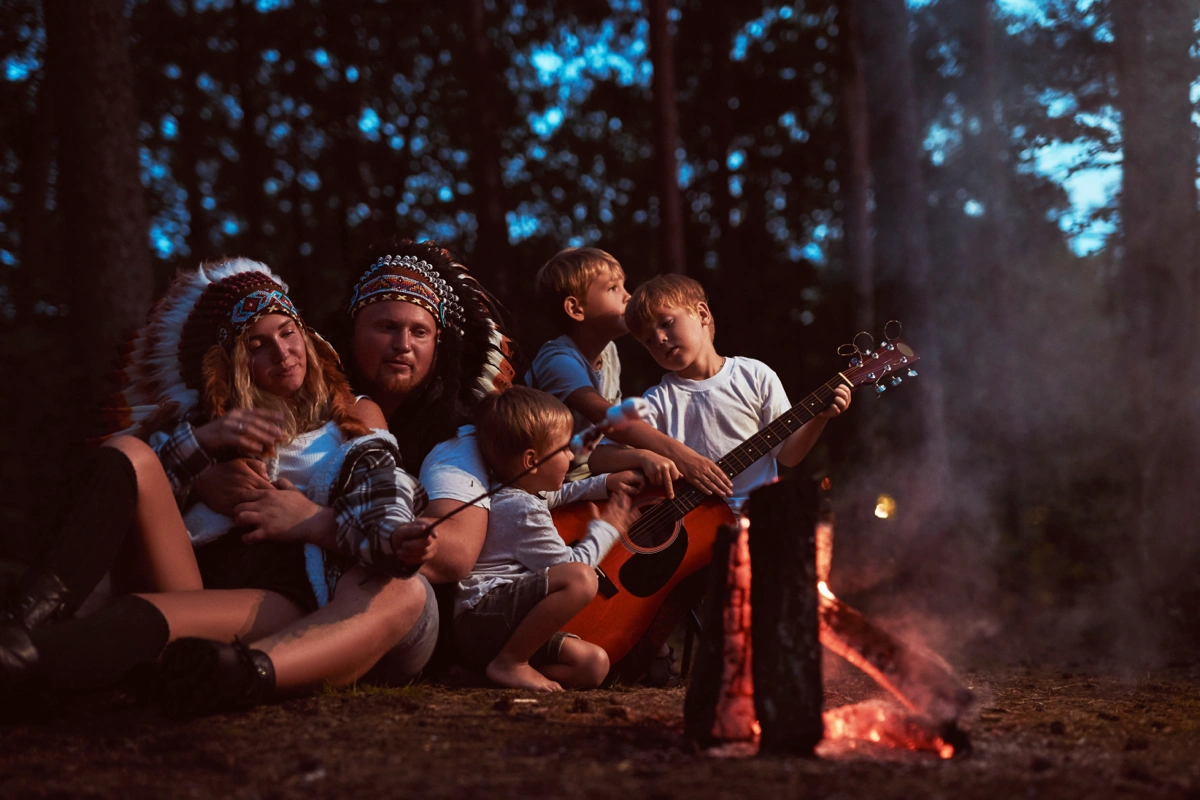Family sitting on the ground around a campfire in a forest at dusk, with two adults wearing feathered headdresses and children roasting marshmallows and playing guitar.