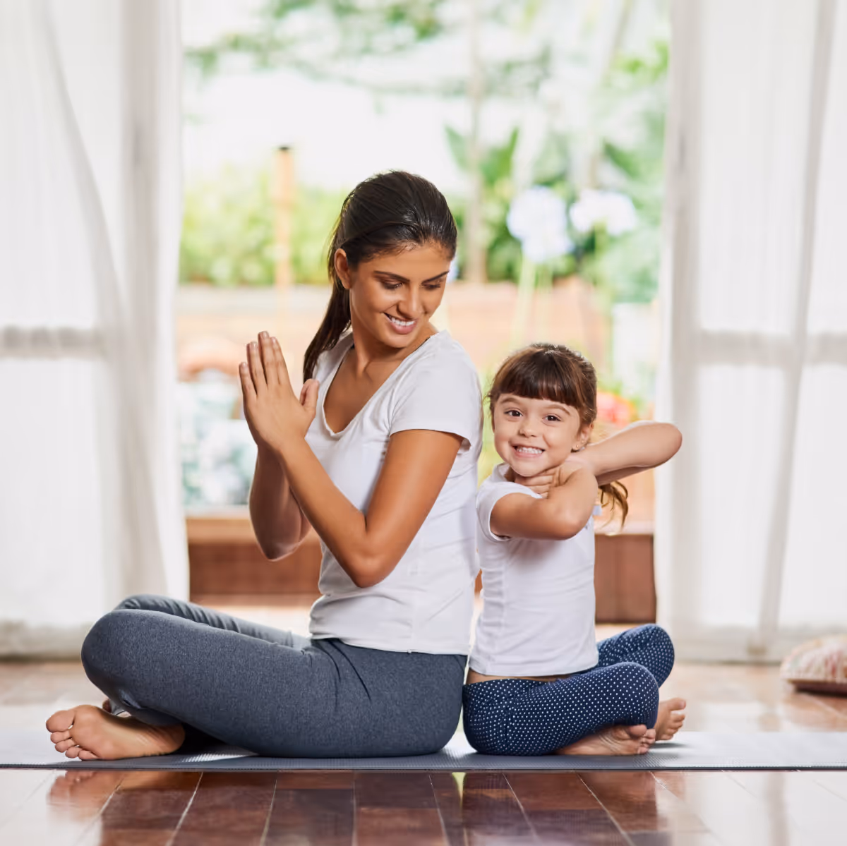 A woman and a young girl sitting back to back on a yoga mat, smiling while practicing yoga indoors.