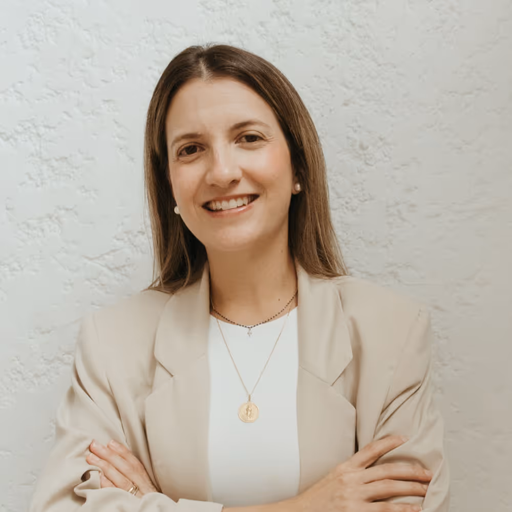 Smiling woman with long brown hair, wearing a beige blazer and white top, standing with arms crossed against a textured light wall.