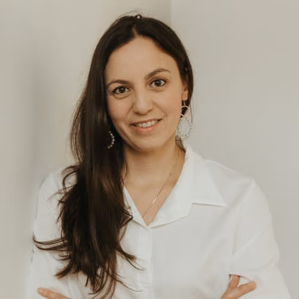 Smiling woman with long dark hair wearing a white blouse and large earrings against a plain background.