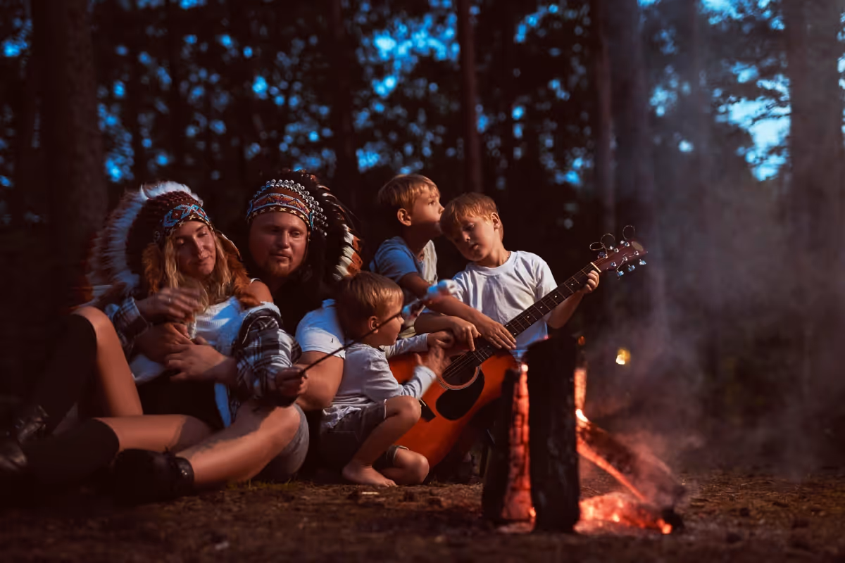 Family sitting by campfire, wearing headdresses and playing guitar at night