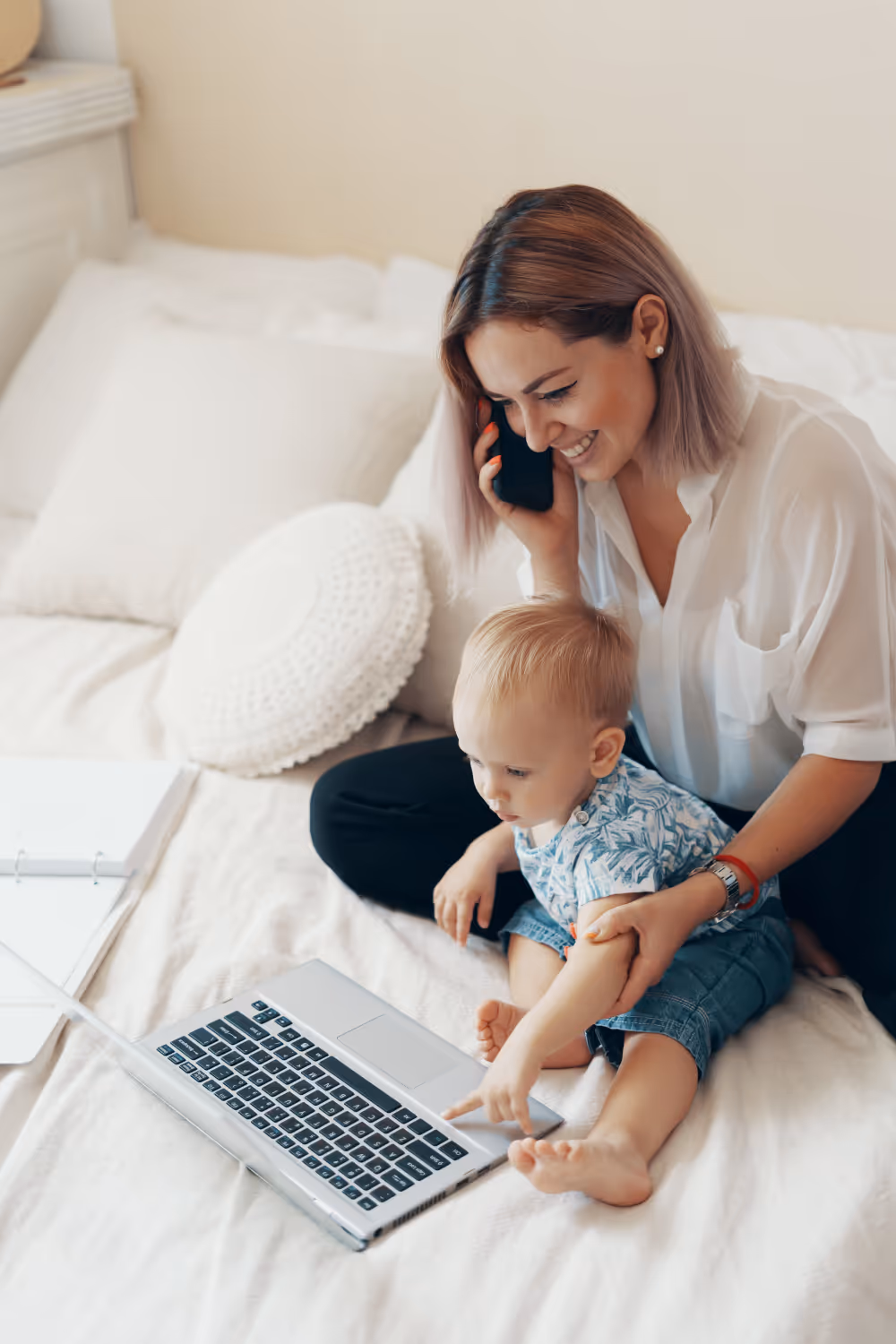 Smiling woman sitting on bed with toddler who is pointing at a laptop keyboard while she talks on the phone.