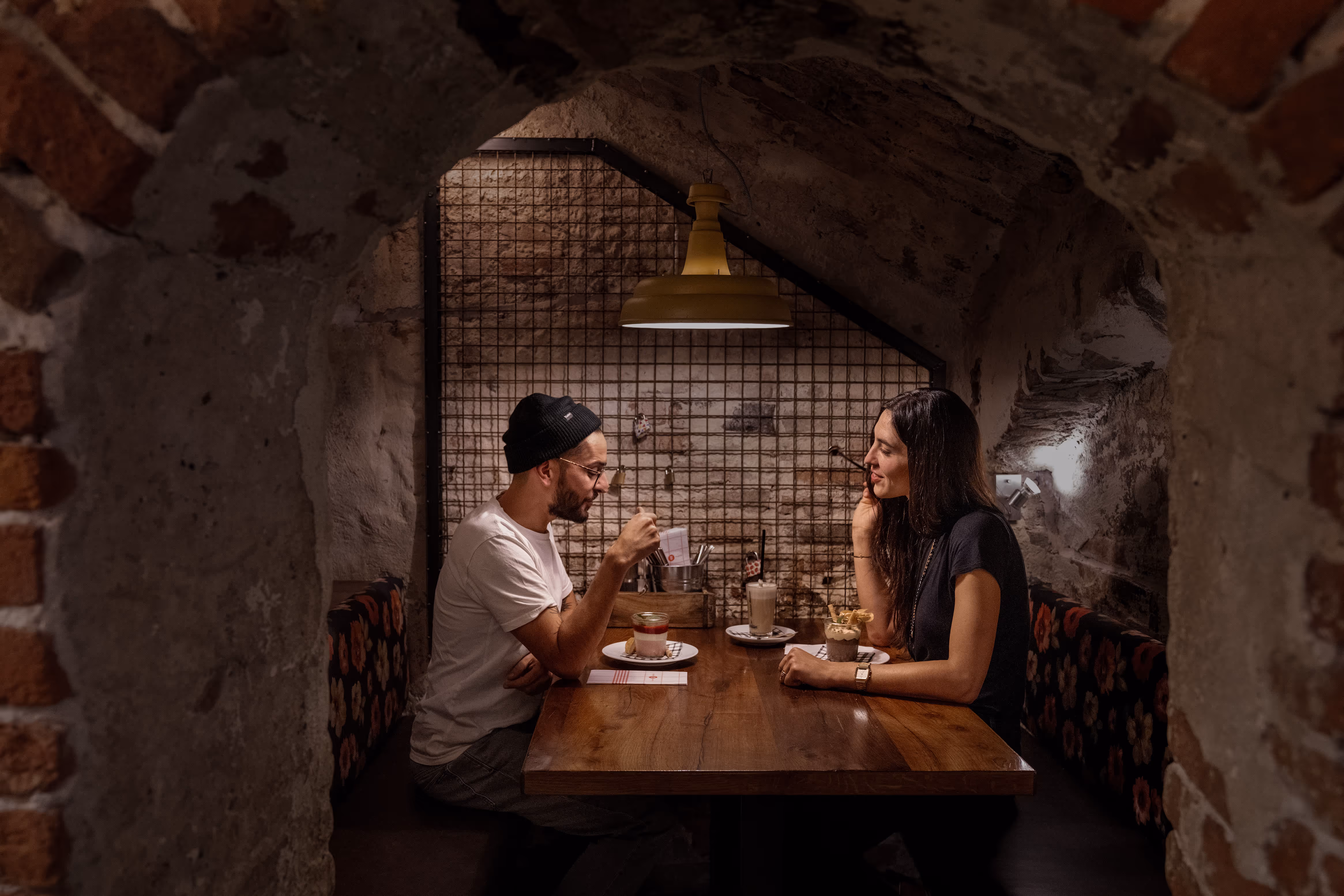Ein Mann und eine Frau sitzen an einem Holztisch in einem gemütlichen, gewölbten Raum mit Backsteinwänden und essen Desserts.