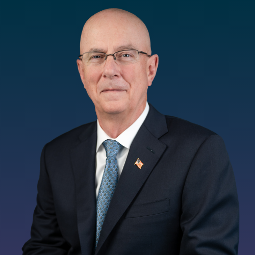 Headshot of male with glasses facing the camera in a dark suite with white dress shirt and blue tie. 