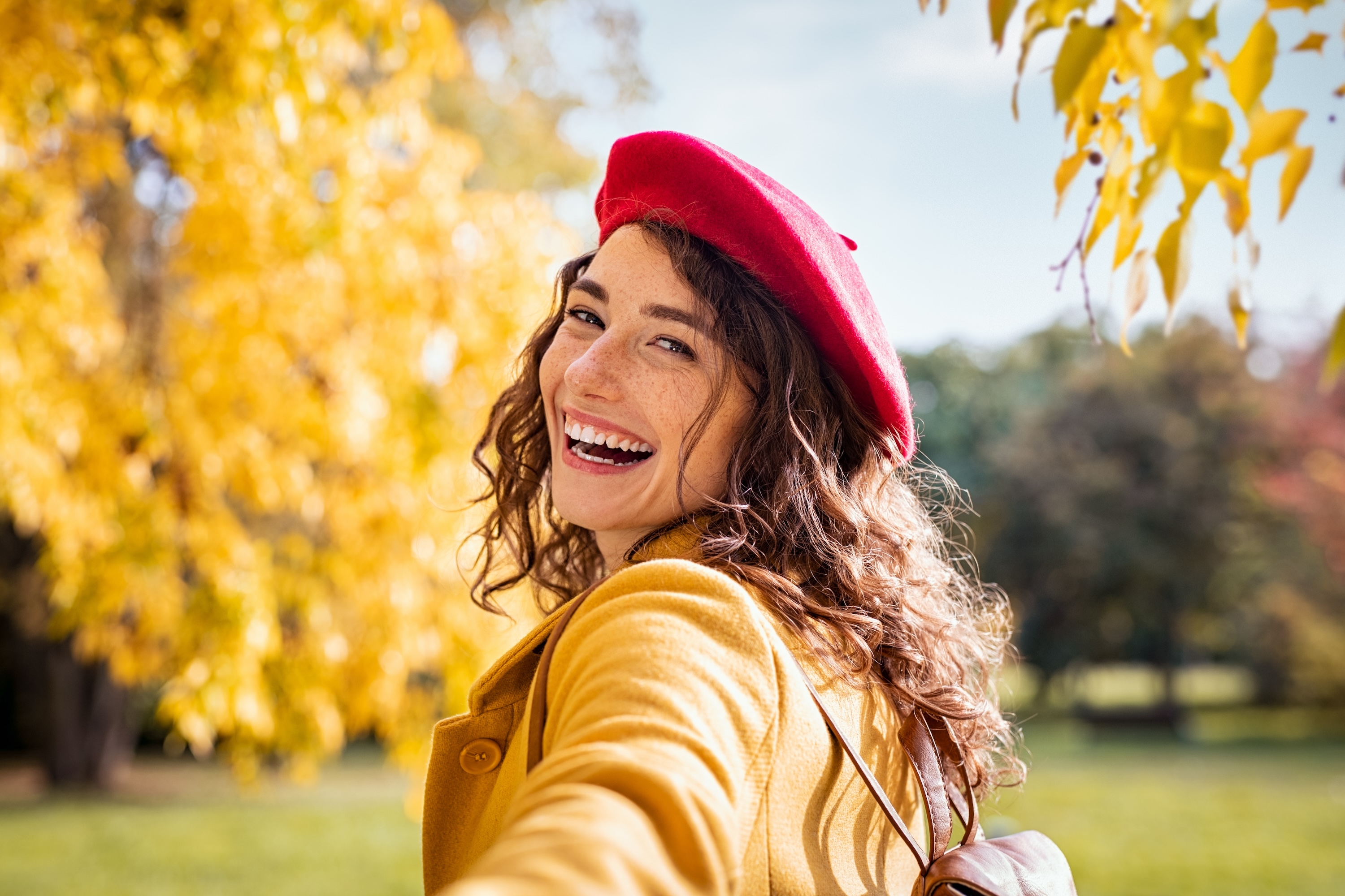 A beautiful, smiling woman in a red beret and yellow coat, enjoying the sunny autumn foliage, representing the scenic outdoor spaces near The Eleanor Daytona Beach in Daytona Beach, FL.