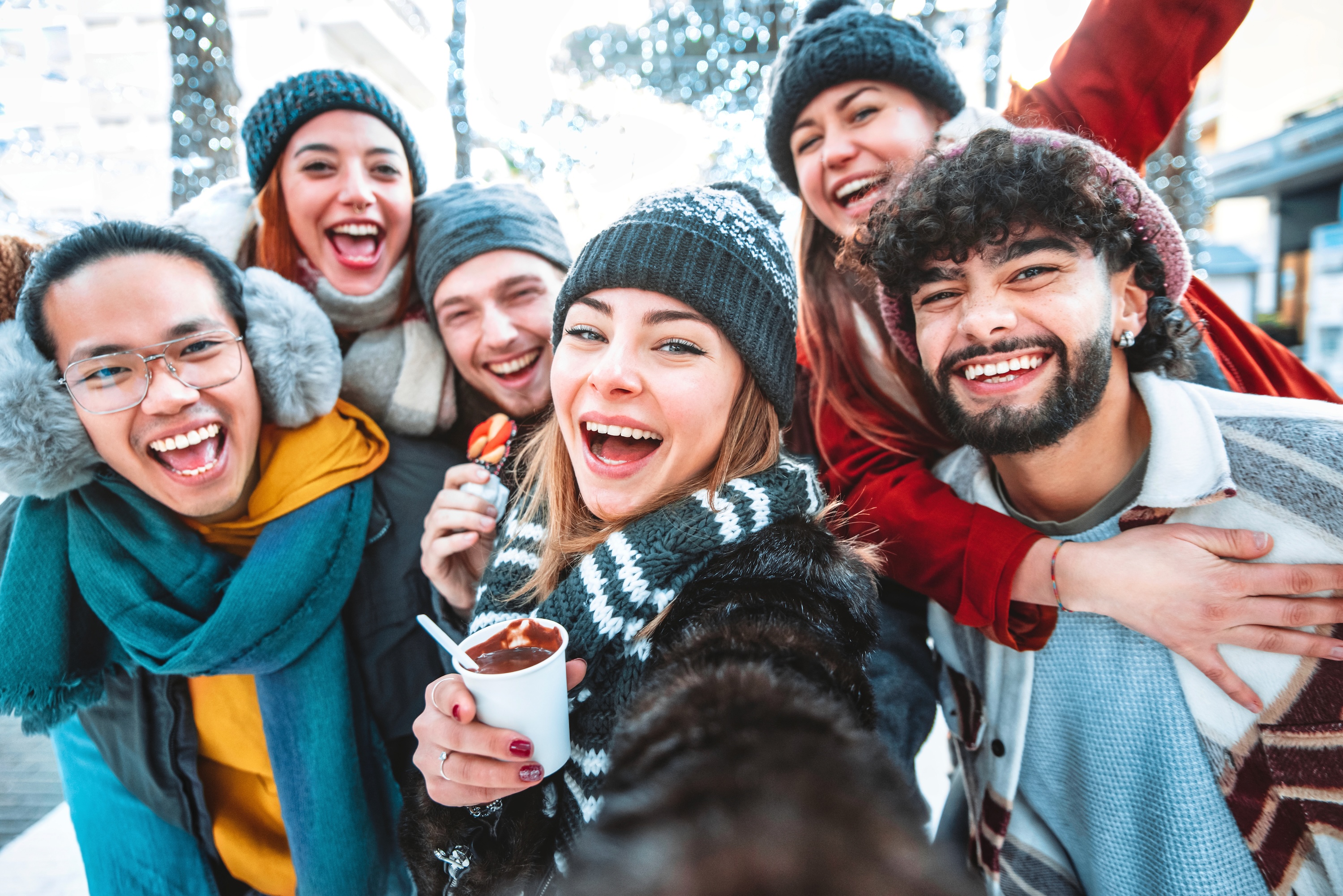 A group of smiling residents posing for a selfie in an outdoor urban setting decorated with festive holiday lights, holding warm beverages near The Eleanor Daytona Beach.