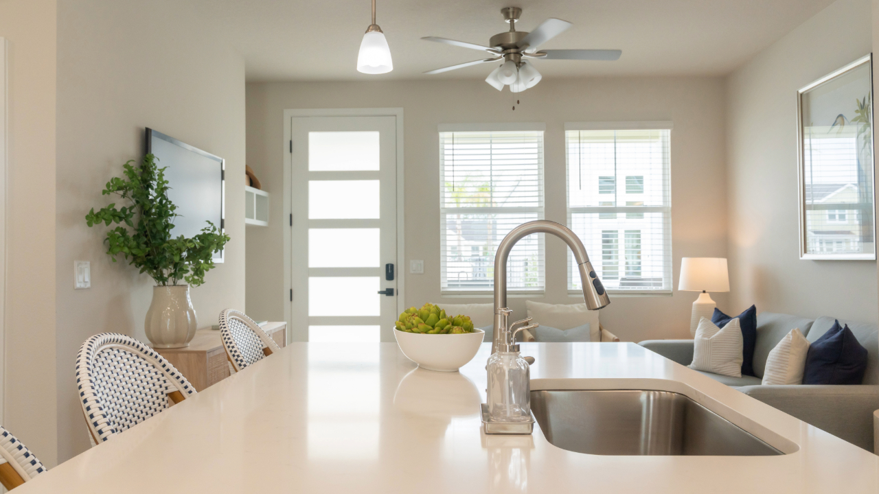 Home living room with a sofa and armchair, with a kitchen featuring white cabinets and stainless steel appliances in the background at The Eleanor Daytona Beach in Daytona Beach, Florida.
