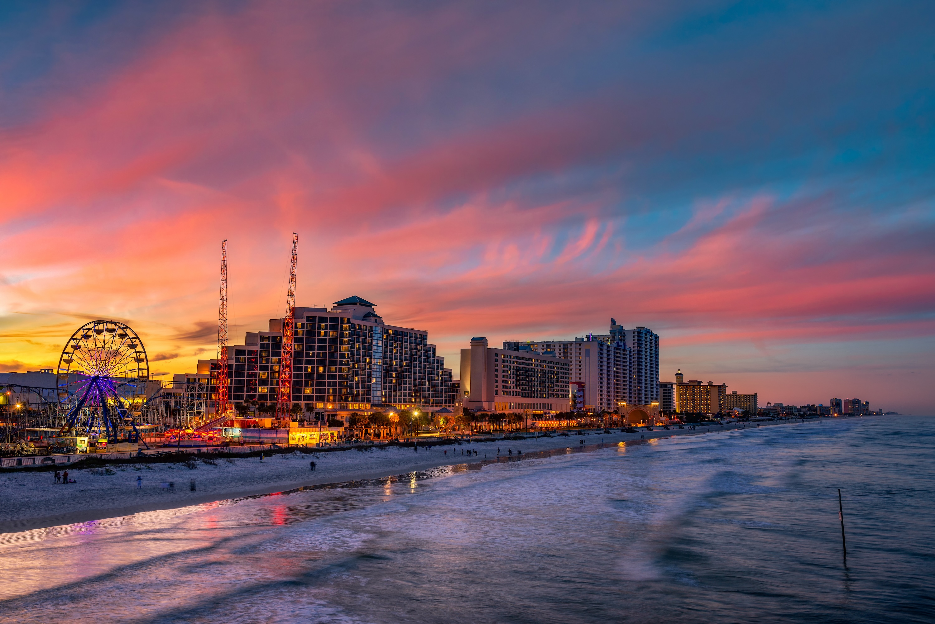 Daytona Beach oceanfront at sunset, with a lit Ferris wheel, boardwalk, and high-rise buildings along the shore