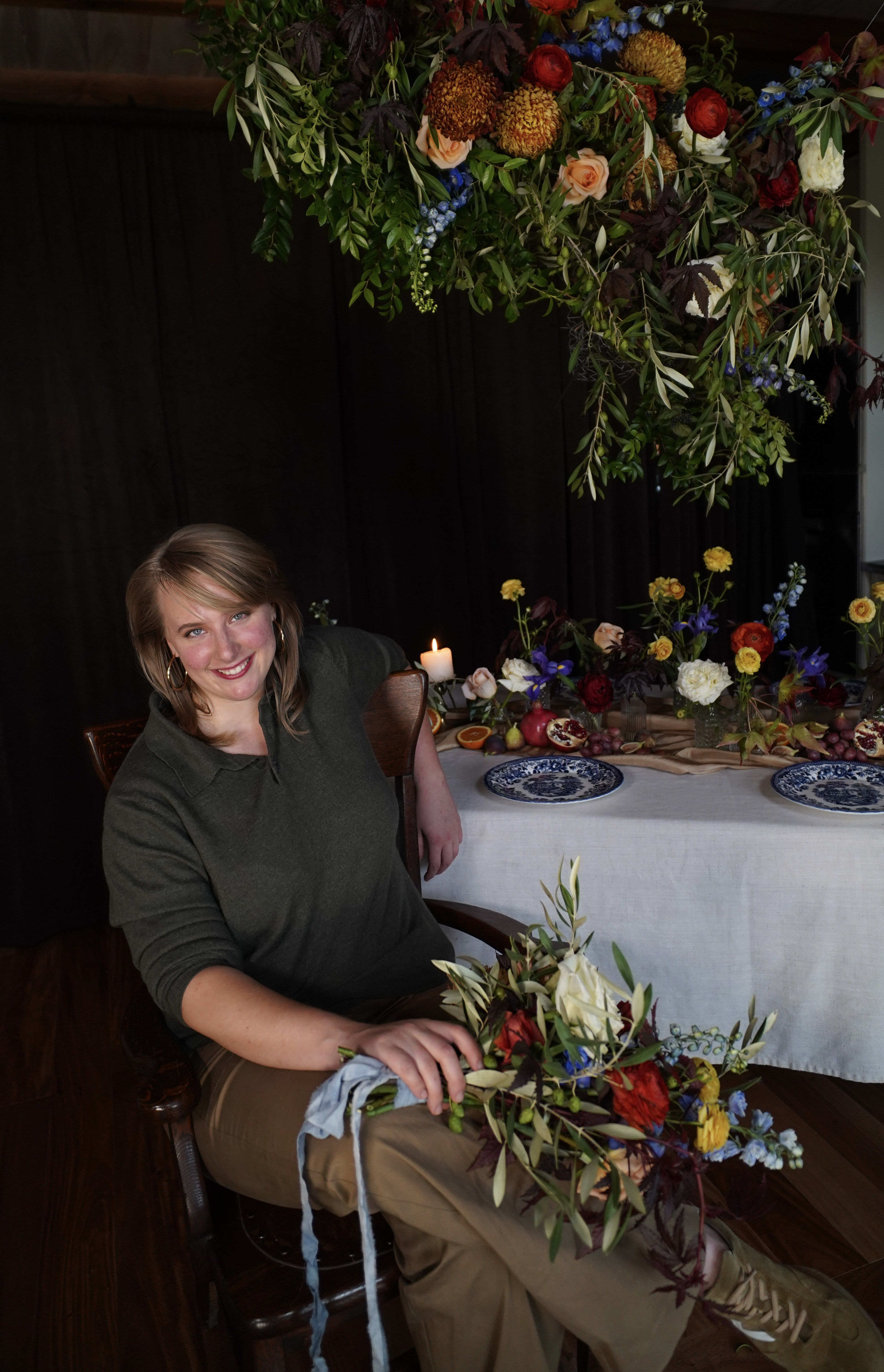 Floral designer and owner of Bluebird & Co., Kaitlyn McDonald, sits at a rich table full of flowers and fruit, set for a dinner, with a colorful and moody hanging floral installation above.