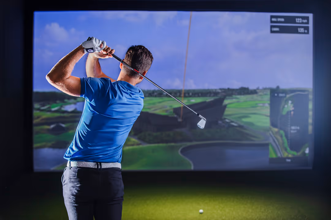 Man in a blue shirt swinging a golf club indoors at a golf simulator with a golf course image on the screen.