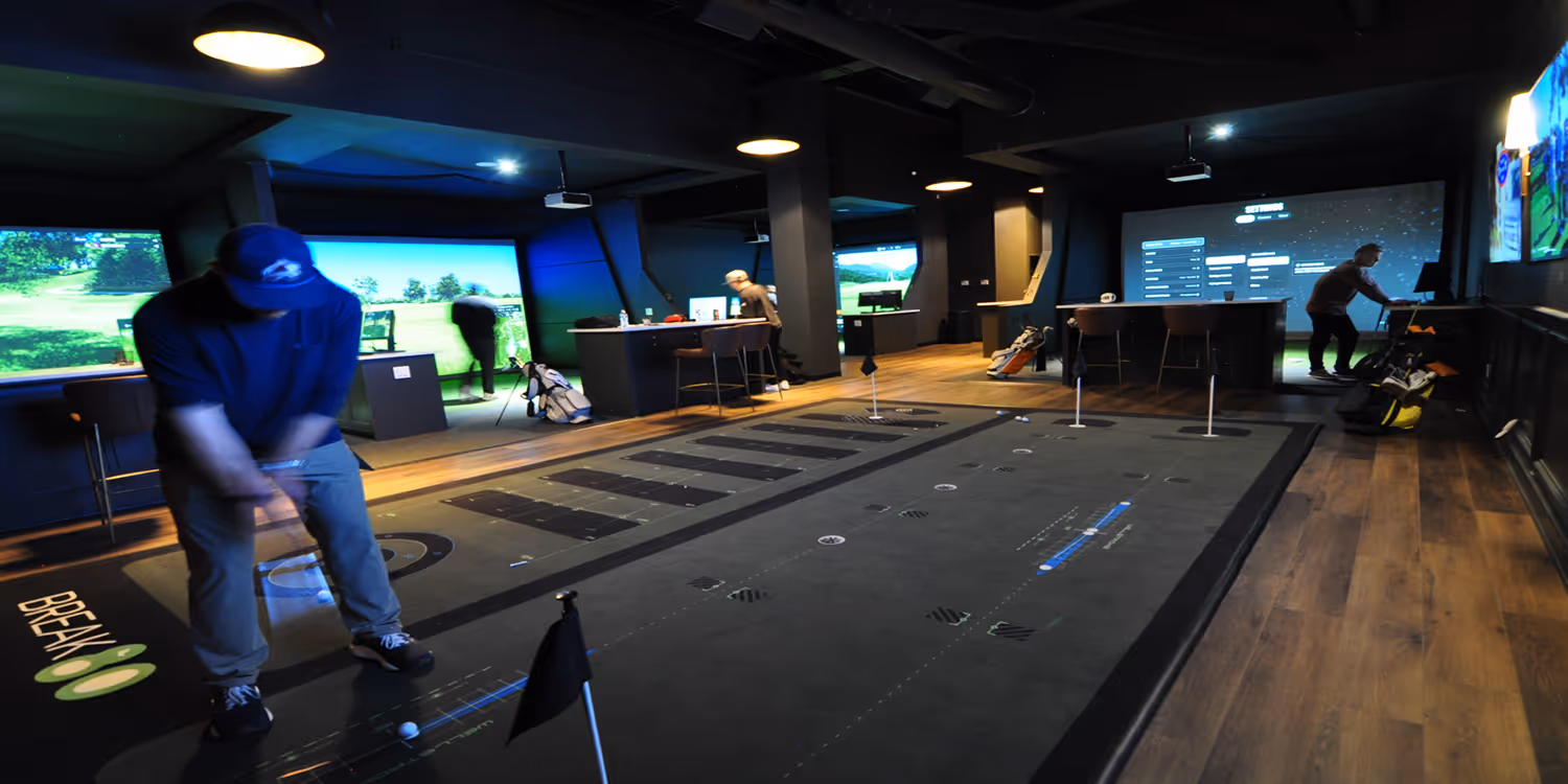 Indoor golf simulator facility with several people playing and practicing golf using virtual screens and putting mats.