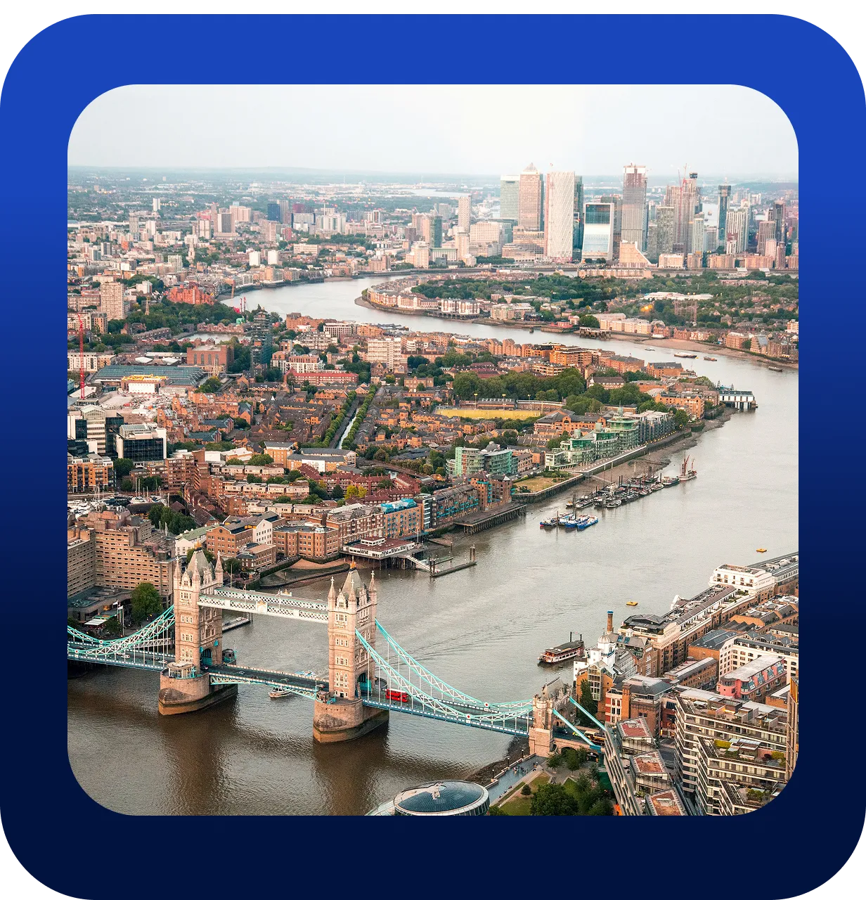 Aerial view of the Tower Bridge crossing the River Thames in London, surrounded by city buildings, residential areas, and the Canary Wharf skyline in the background.