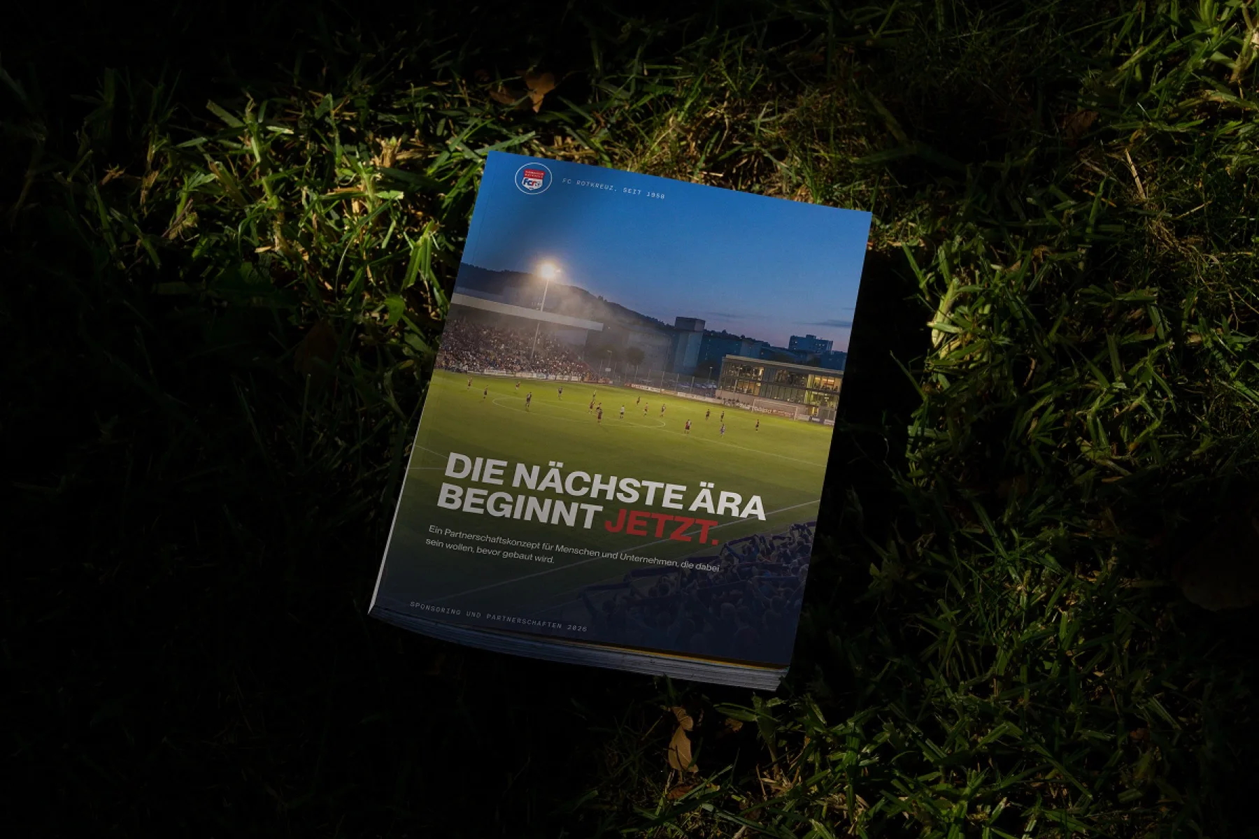 The printed FC Rotkreuz sponsoring dossier lying on grass at night, its cover showing a floodlit stadium and the headline Die naechste Aera beginnt jetzt