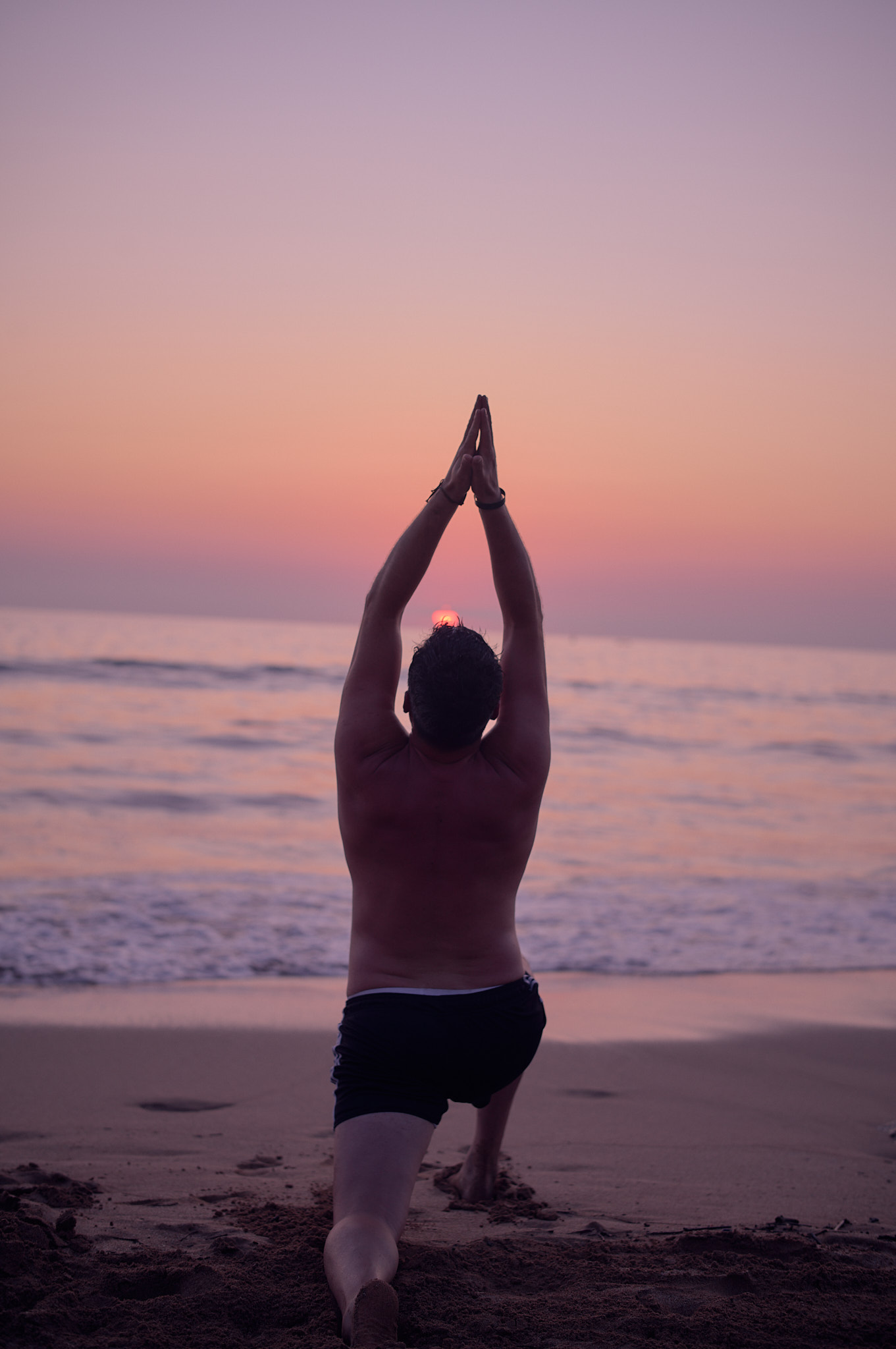 Trainee in standing salutation viewed from behind on the wet sand as the sun sets over the Pacific.