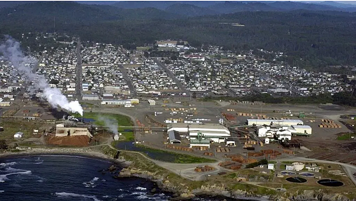 Aerial view of a coastal town with industrial buildings, stacks emitting white smoke, residential area in the background, and the ocean in the foreground.