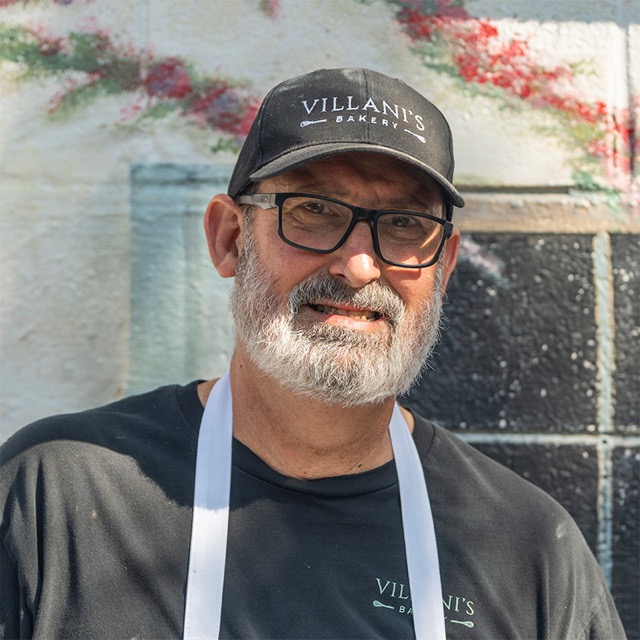 Paul Villani, co-owner of Villani’s Bakery, wearing a Villani’s Bakery cap, black shirt with the bakery logo, white apron, and glasses, standing in front of a painted wall mural.
