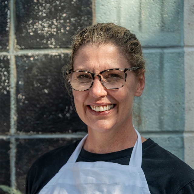Amanda Villani, co-owner of Villani’s Bakery, smiling while wearing glasses, a white apron, and a black shirt, standing in front of a painted cinder block wall.