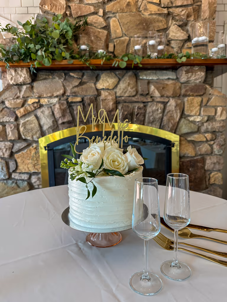 White wedding cake with roses and gold topper, set in front of a stone fireplace — by Villani’s Bakery.