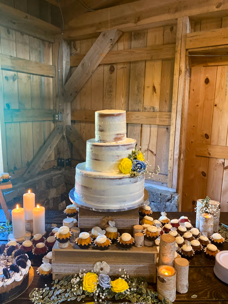 Three-tier rustic wedding cake with cupcakes and candles in barn setting by Villani's Bakery