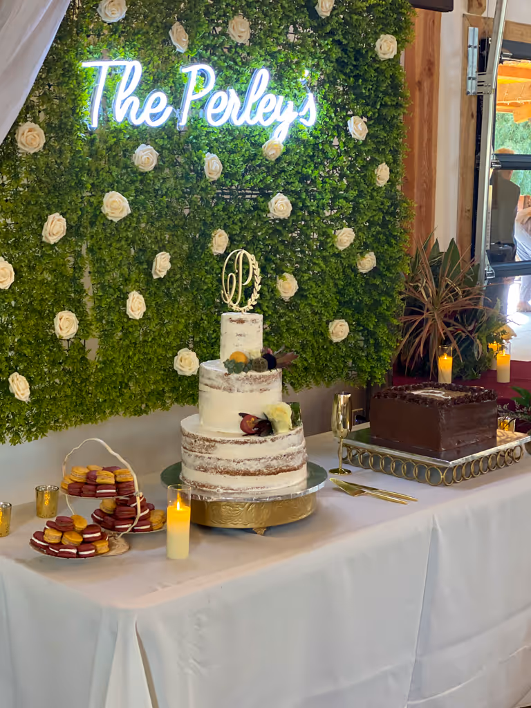 Wedding dessert display with semi-naked cake and groom's cake against floral wall by Villani's Bakery