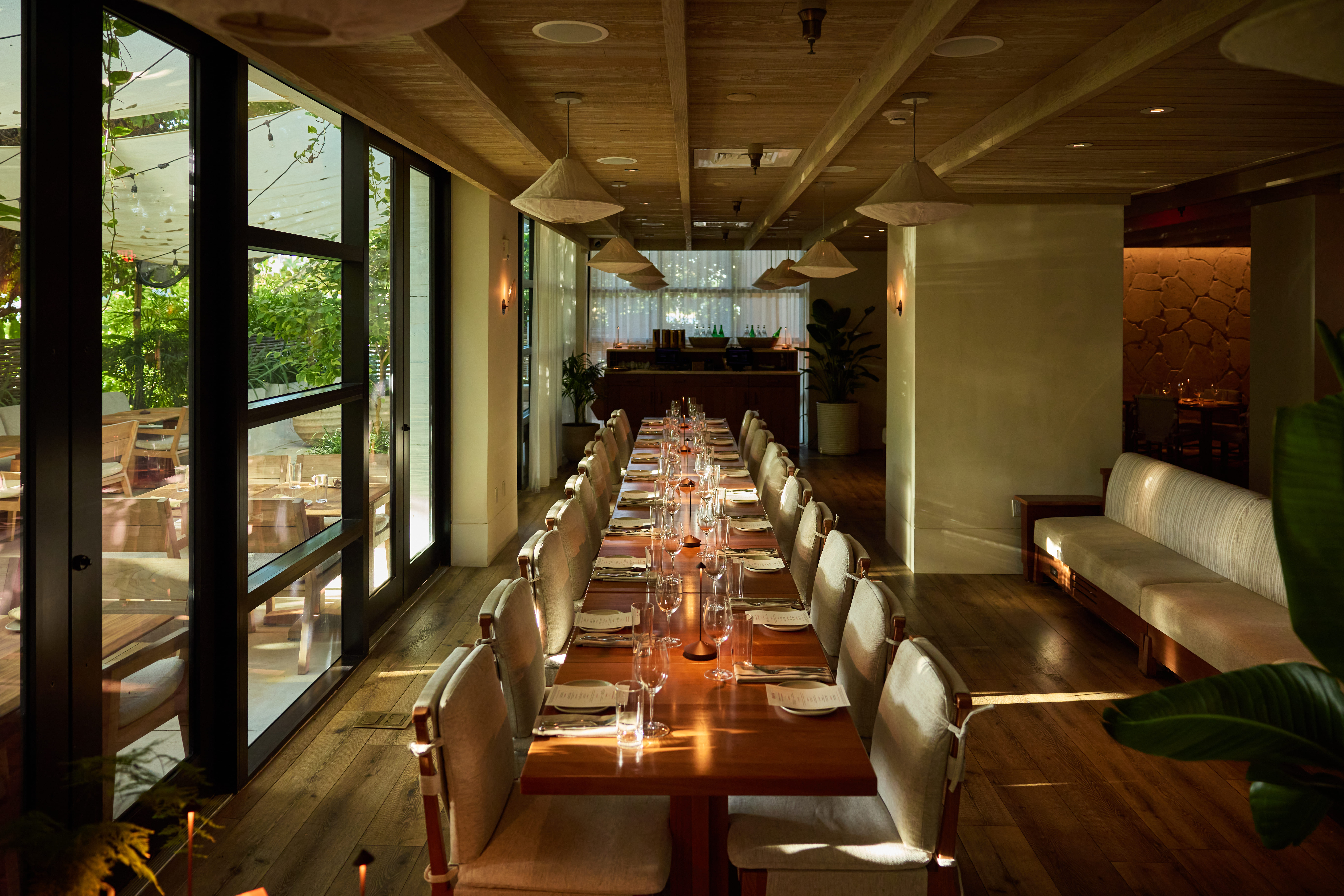 Long communal dining table set for dinner inside AVIV, surrounded by warm wood finishes and floor-to-ceiling windows overlooking a lush patio.