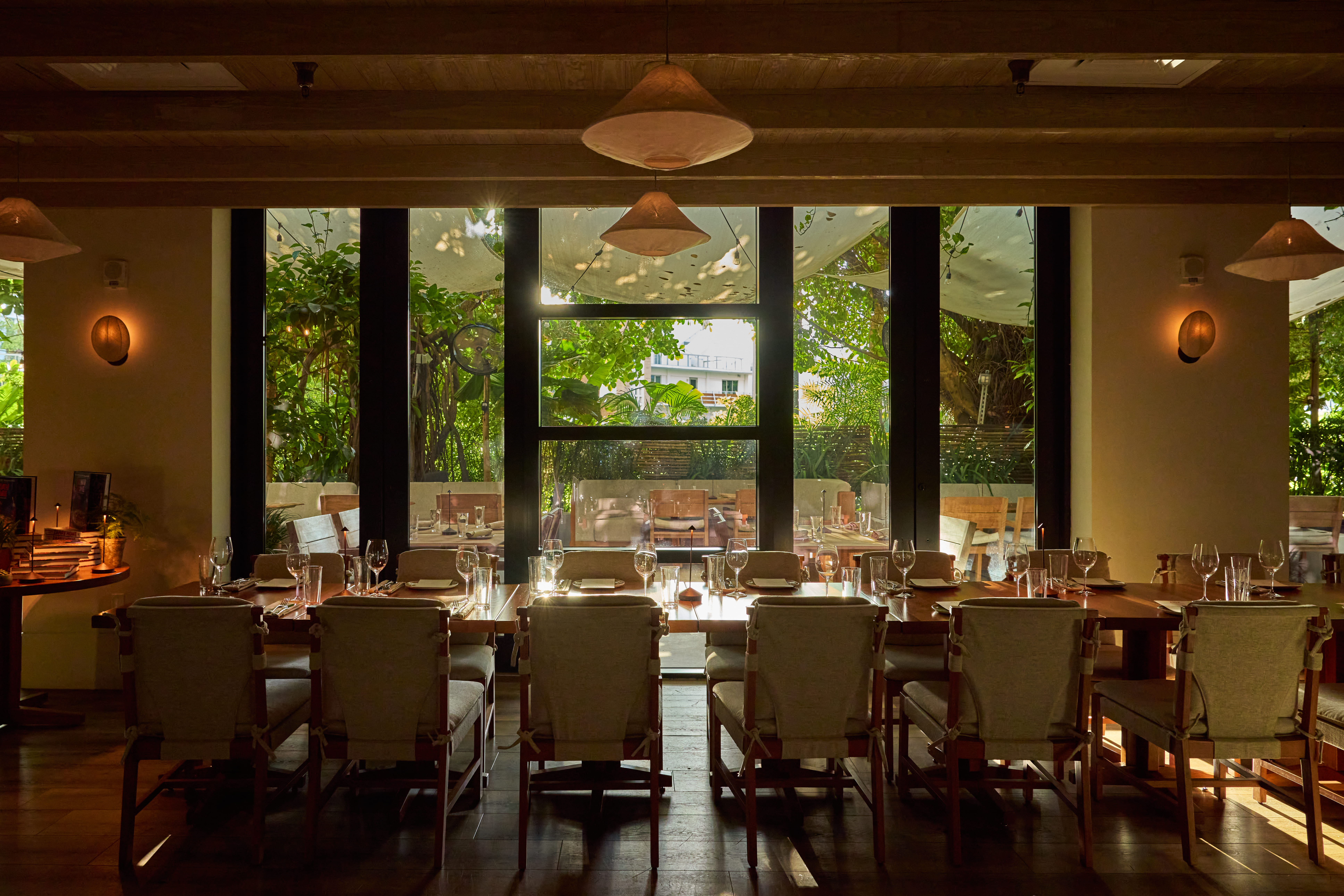 Sunlit dining room at AVIV with a long set table, pendant lighting and large windows overlooking the garden patio.