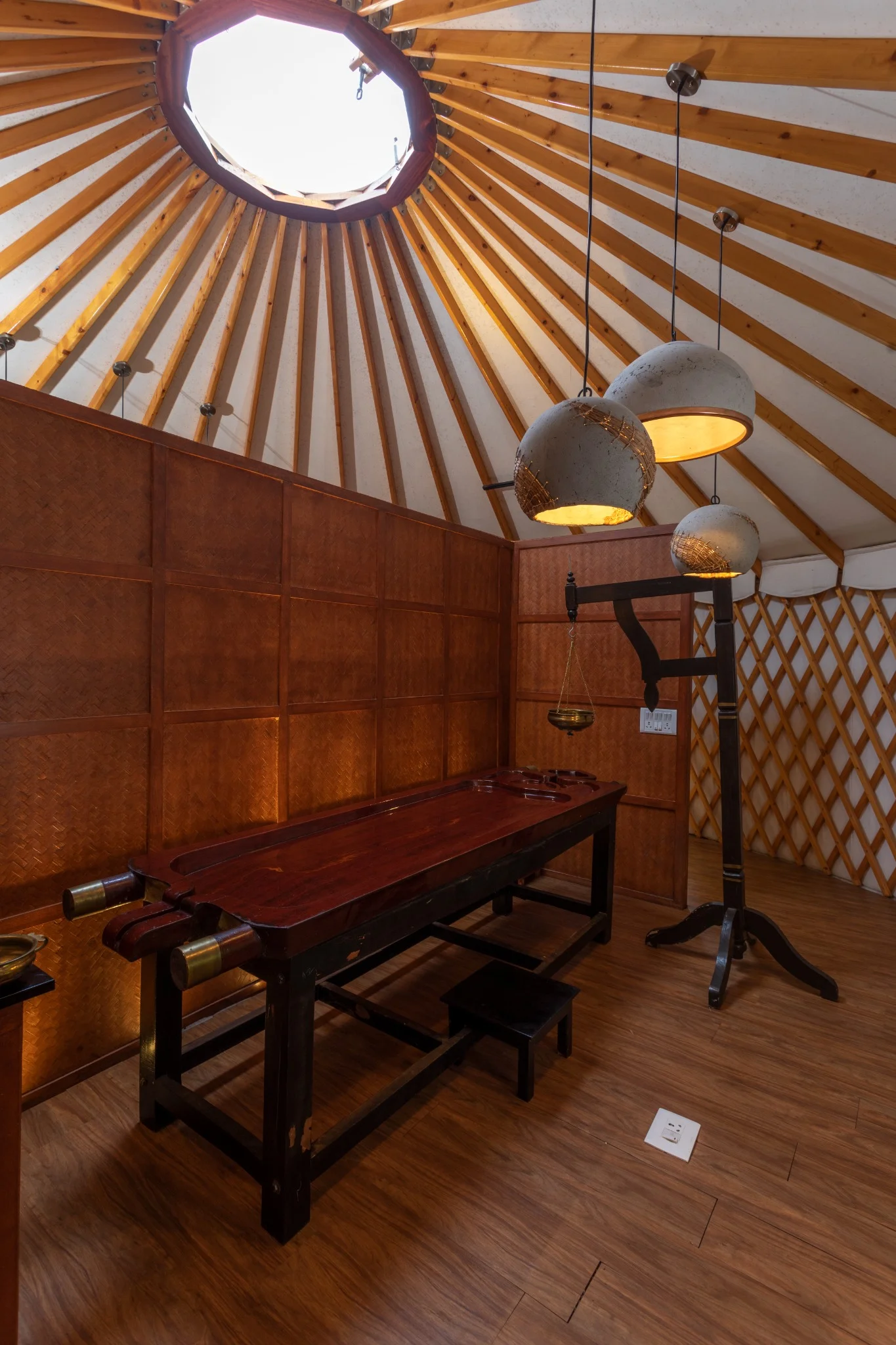 Interior of a yurt with a traditional wooden Ayurvedic massage table under hanging pendant lights, featuring wooden panel walls and a skylight.