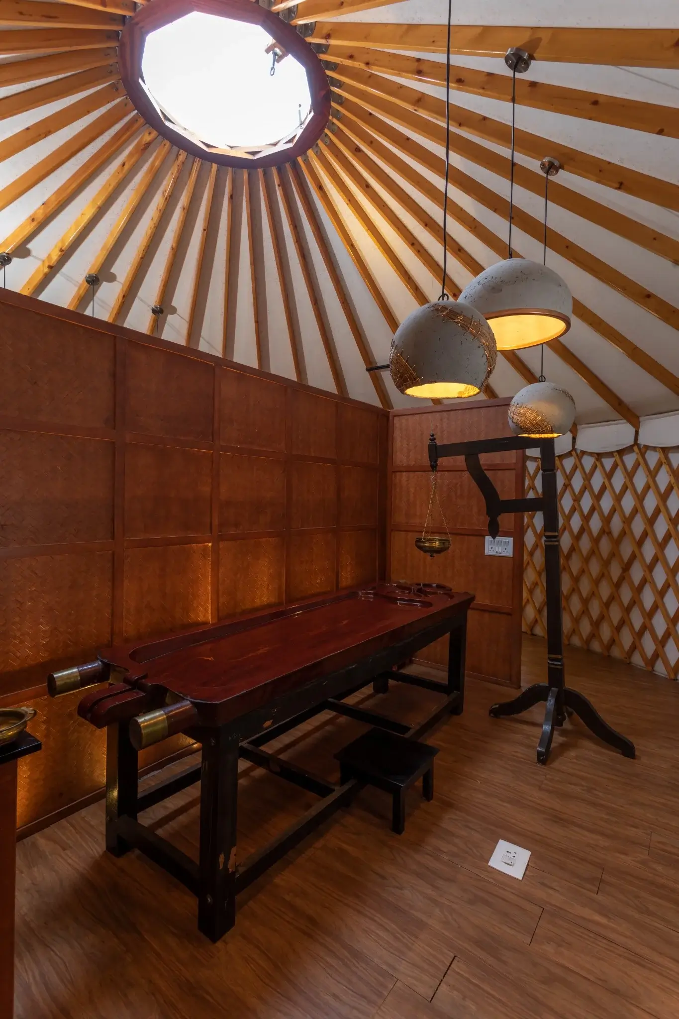 Interior of a yurt with a traditional wooden Ayurvedic massage table under hanging pendant lights, featuring wooden panel walls and a skylight.