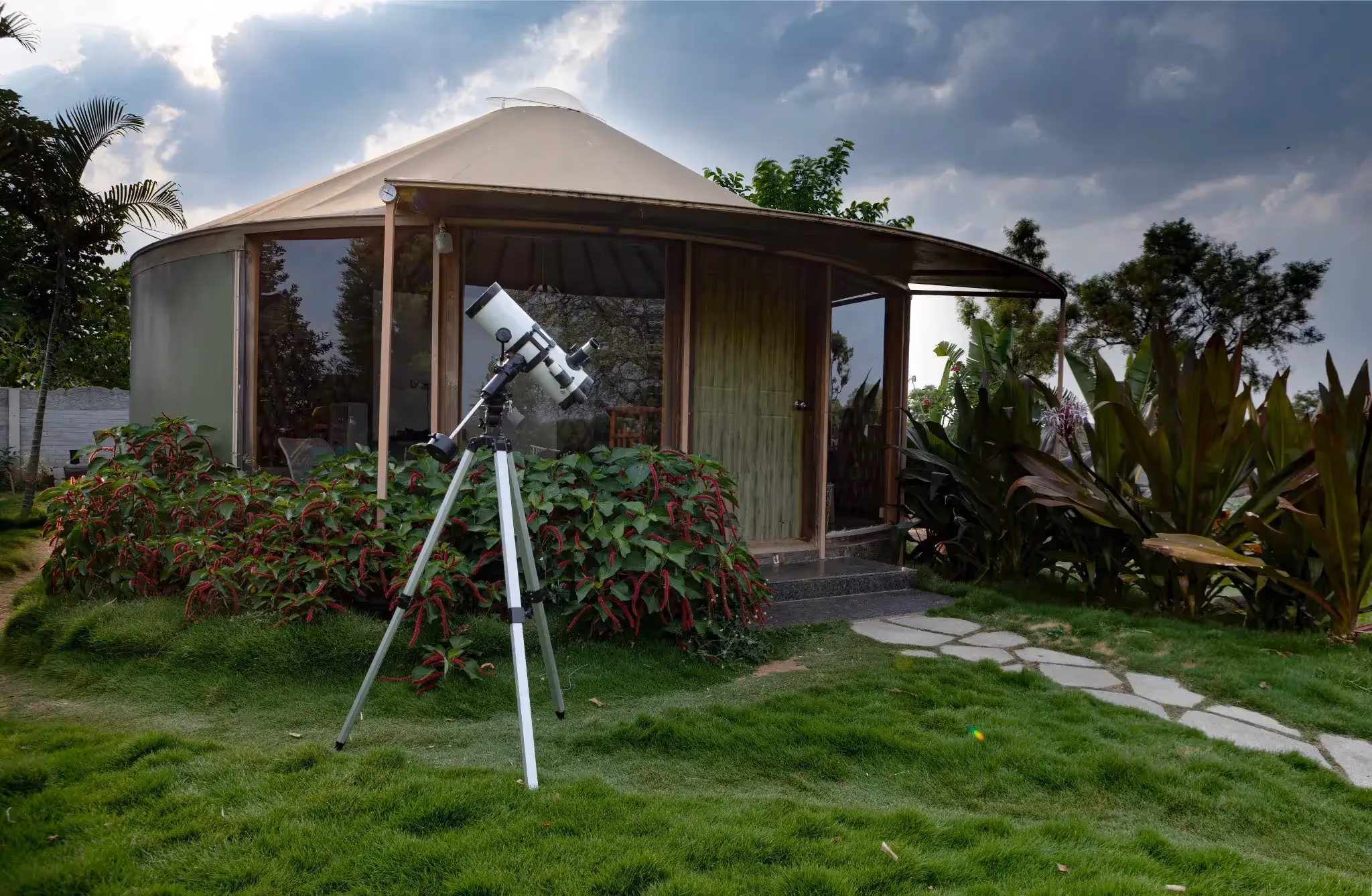 Luxury glamping yurt with a telescope outside, surrounded by vibrant greenery and dramatic sky backdrop.