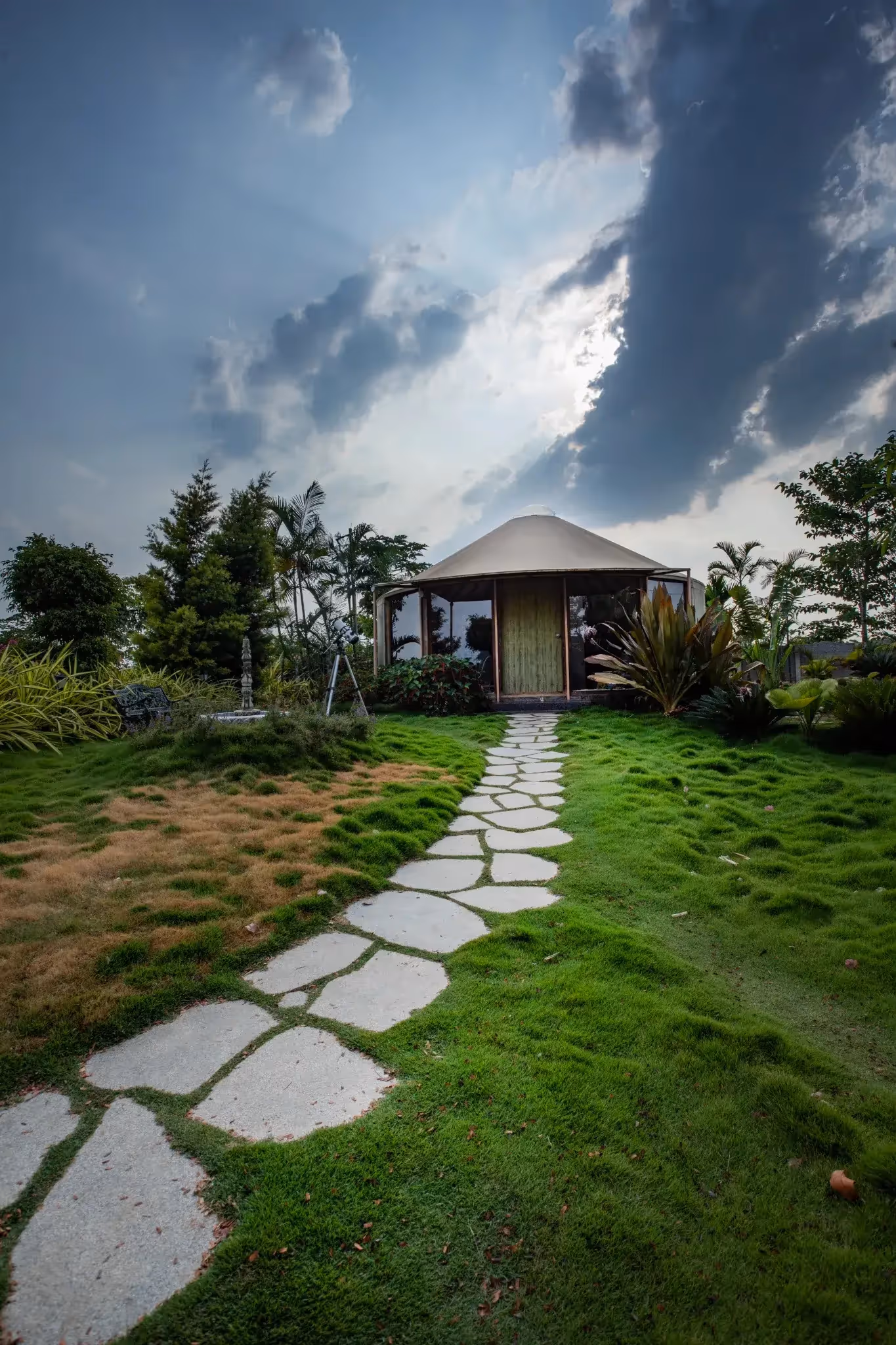 Eco-friendly yurt nestled in lush greenery with a stone path leading to the entrance under dramatic skies.