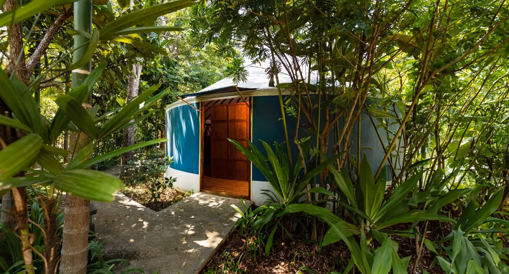 Blue and white yurt with wooden door partially open, surrounded by tropical foliage.