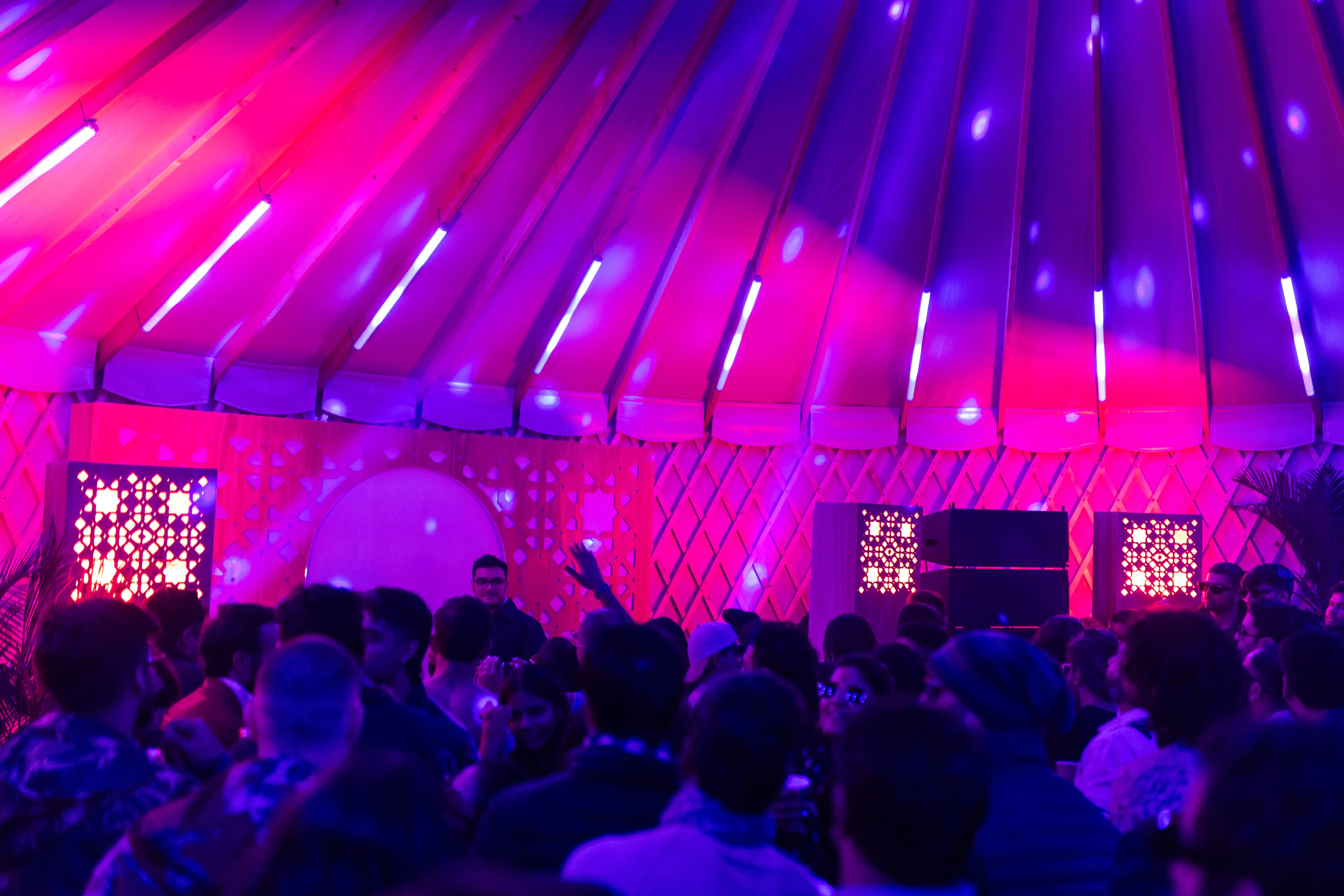 Crowded yurts interior during party with purple neon lighting, lattice walls, and dancing guests inside