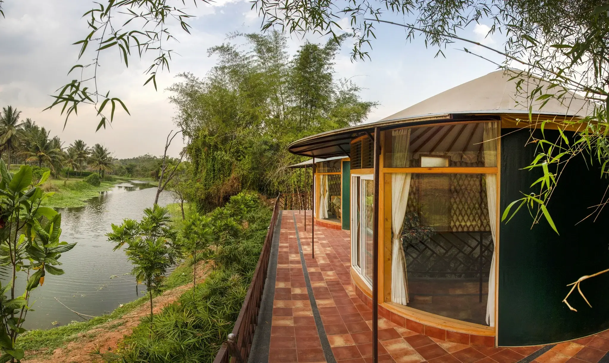 Riverside resort yurts with large windows and tiled terrace, surrounded by bamboo, palms, and lush greenery