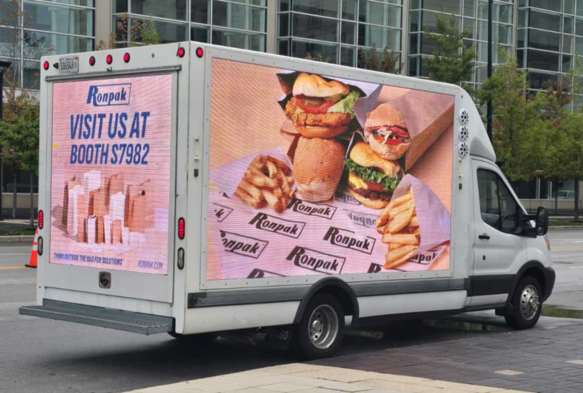 White delivery truck with digital screens displaying Ronpak advertisement featuring burgers, fries, and a message to visit booth S7982.