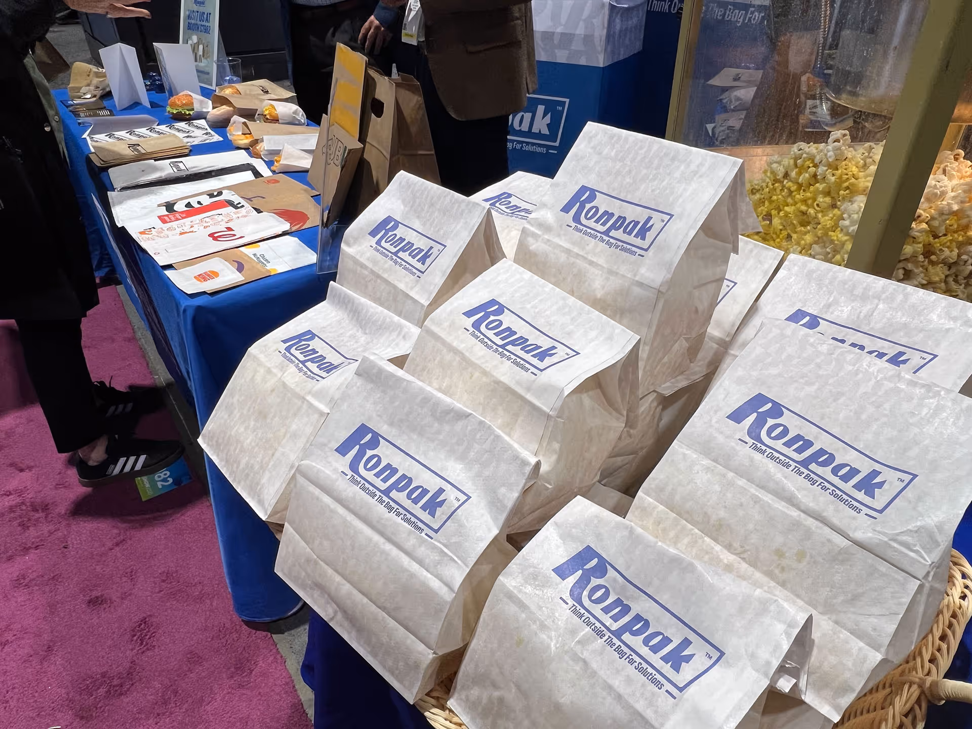 Display of white folded Ronpak paper bags on a table with various other food packaging items in the background and a popcorn machine.