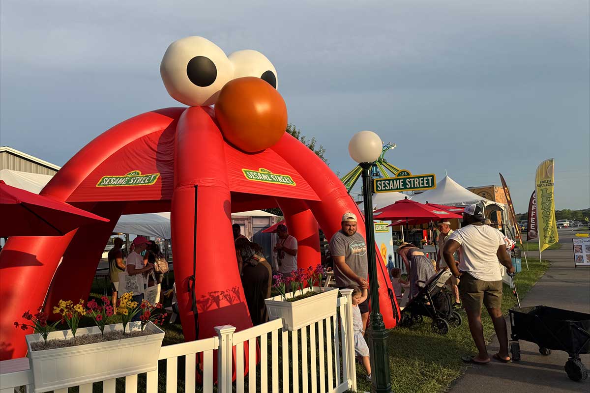 Outdoor event with a large inflatable red Sesame Street character arch, people walking and standing around, and a street sign reading 'Sesame Street'.