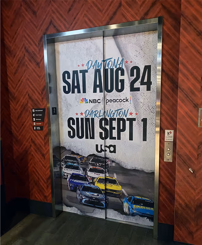 Elevator doors decorated with a racing-themed advertisement showing NASCAR cars and race dates: Daytona on Saturday, August 24, and Darlington on Sunday, September 1, with NBC, Peacock, and USA Network logos.