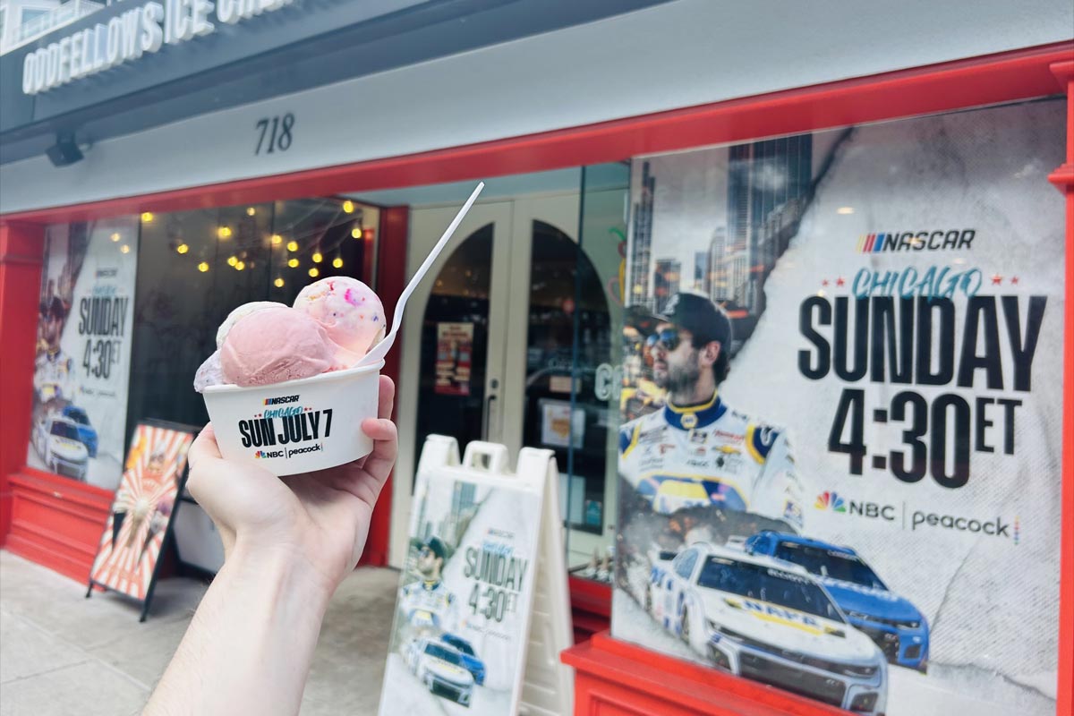 Hand holding a cup of pink and multicolor ice cream with a spoon in front of a red storefront displaying posters and banners promoting a NASCAR Chicago event airing Sunday at 4:30 ET on NBC and Peacock.