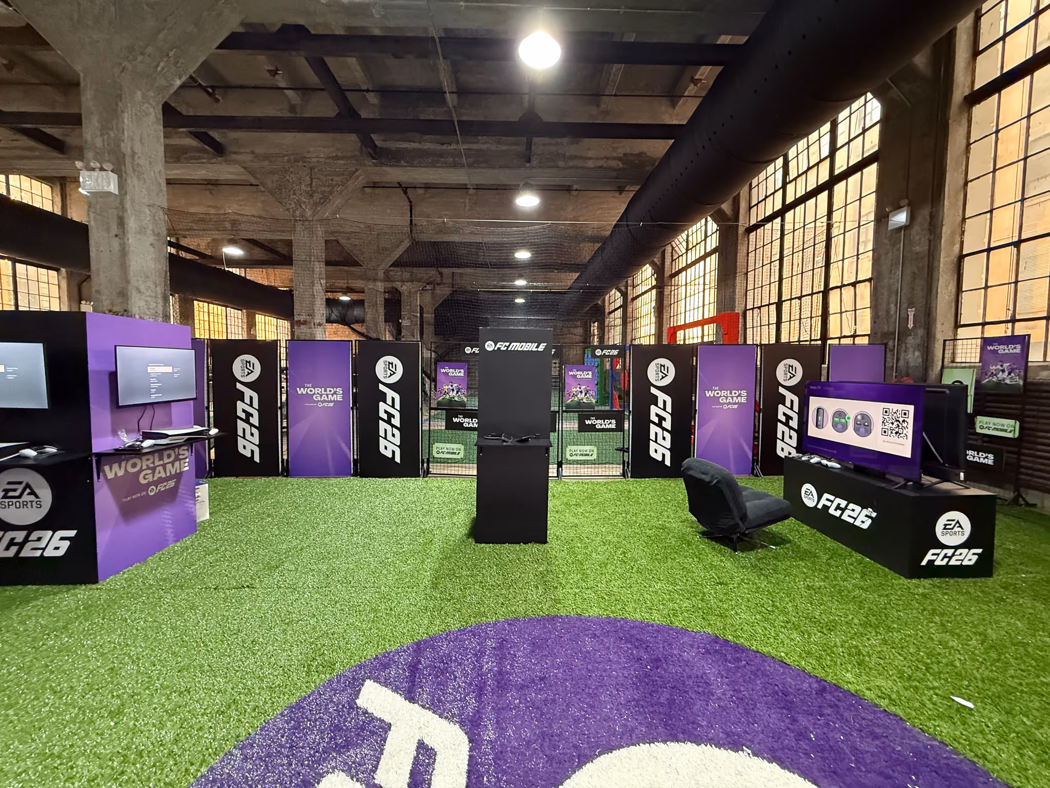 Indoor setup of a gaming event space with artificial green turf flooring and a large purple circle with white EA Sports FC26 logo in foreground, multiple screens and branding panels with EA Sports FC26 and World’s Game logos arranged along walls and center, black gaming chair and central kiosk under industrial ceiling and large windows.