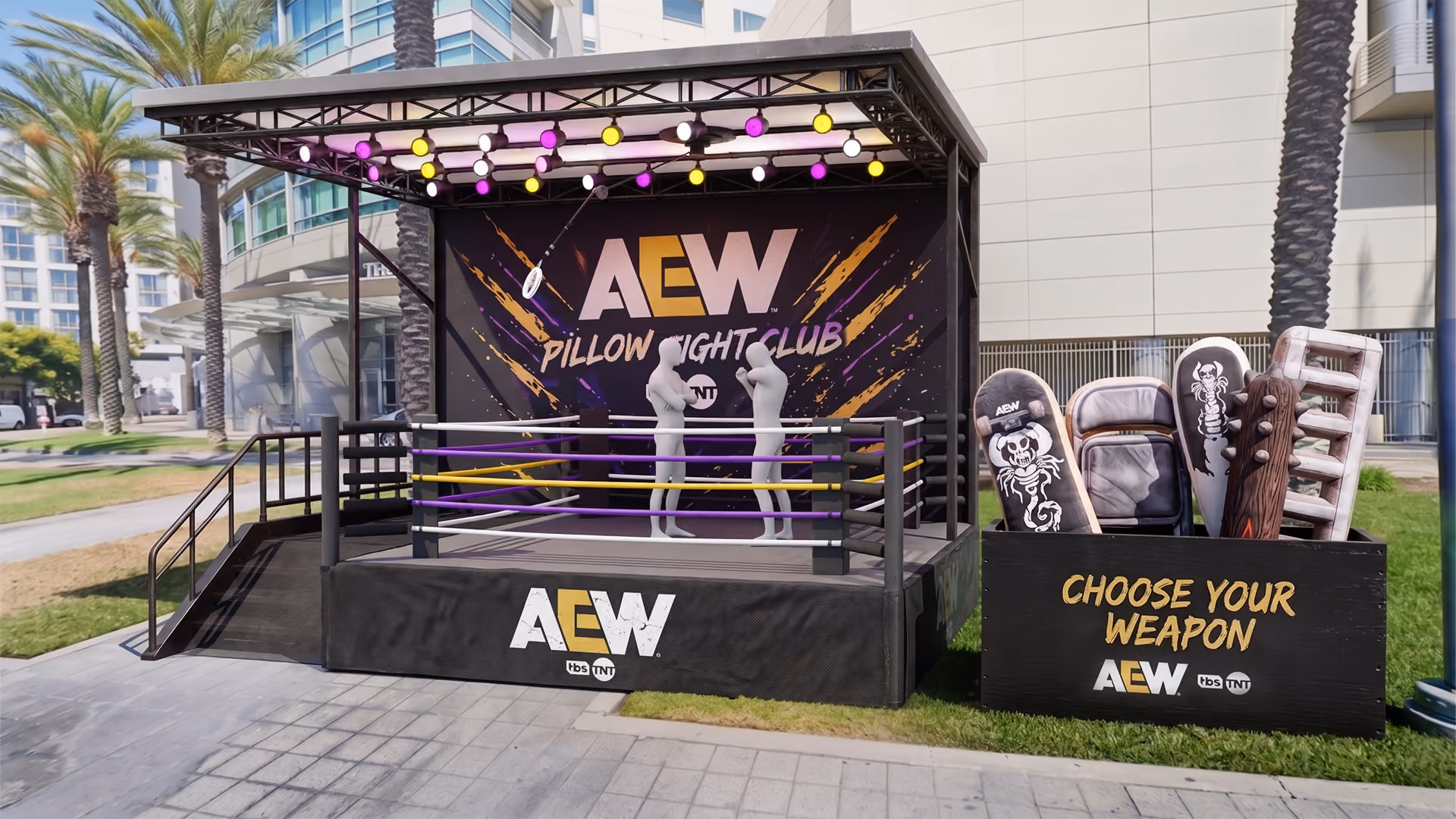 Outdoor setup featuring a black wrestling ring with purple and yellow ropes under a roof with purple, yellow, and white lights, and a backdrop reading 'AEW Pillow Fight Club TNT'. Next to the ring is a black box with various pillows designed as weapons and a sign that says 'Choose Your Weapon AEW TNT'.