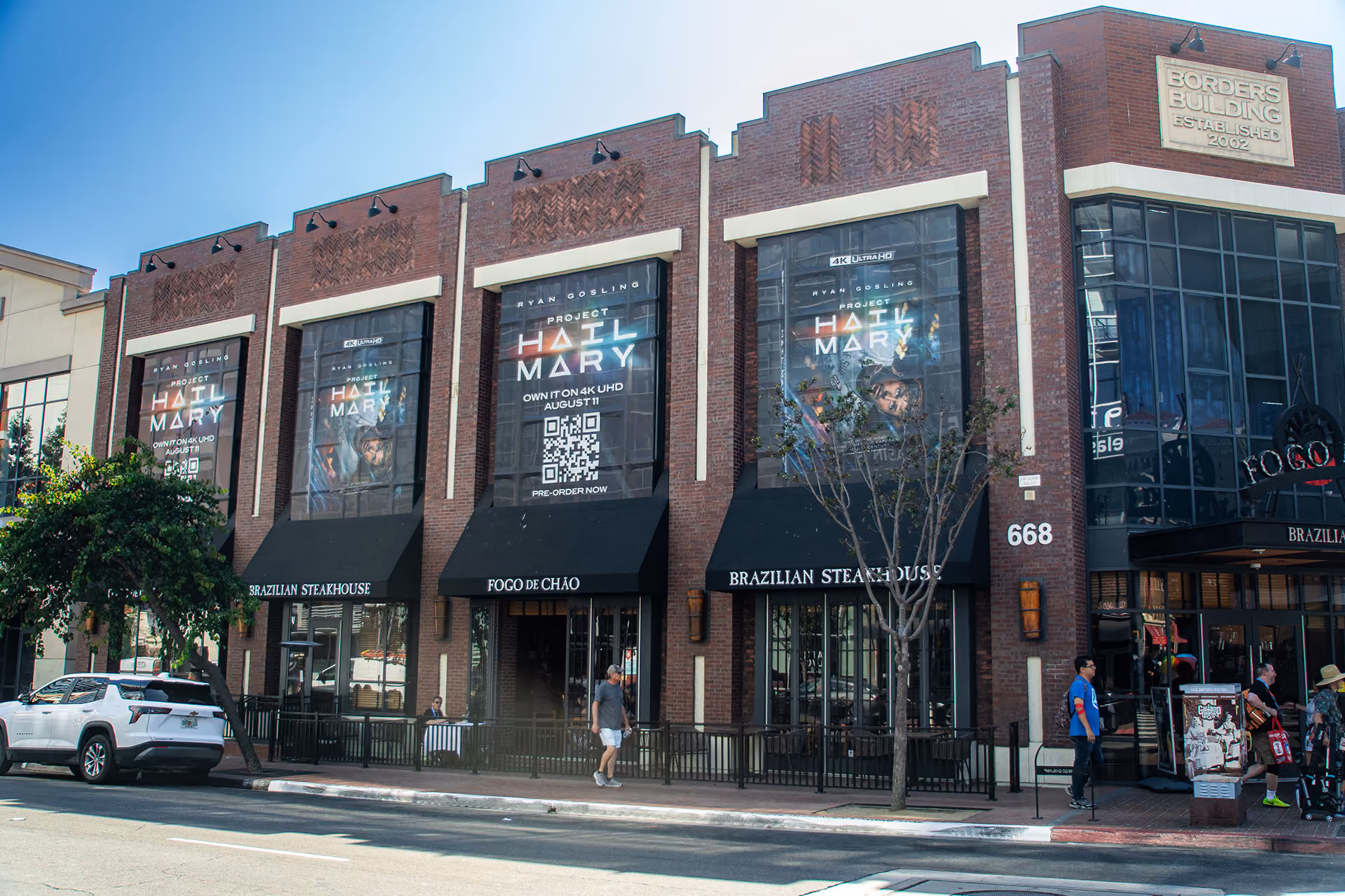 Brick building with large black awnings and tall windows displaying posters for the movie 'Project Hail Mary,' featuring Ryan Gosling, with storefront signage for Fogo de Chão Brazilian Steakhouse and people walking on the sidewalk.